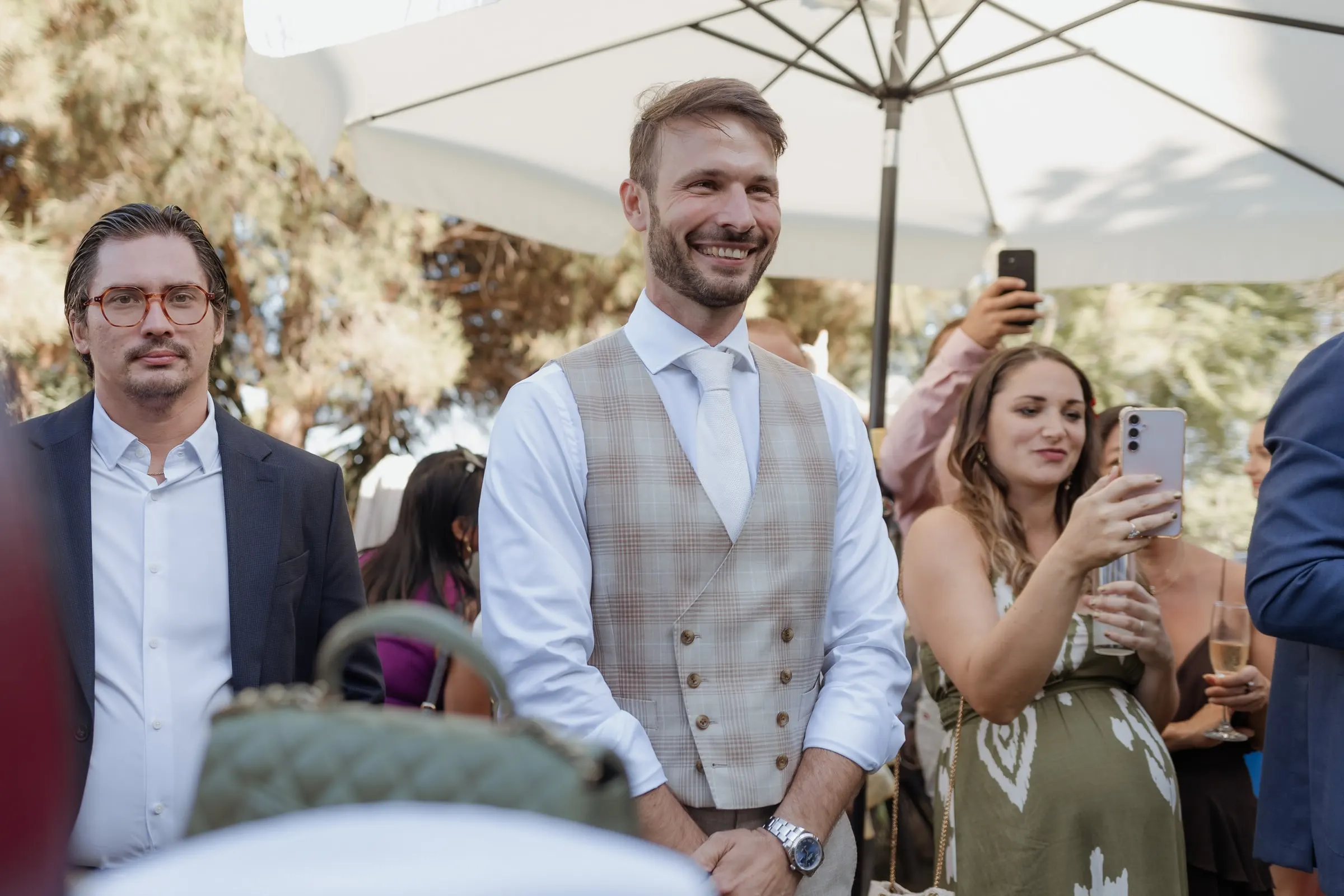 The groom in a tweed waistcoat smiling as guests film on their phones, Hacienda San José, Mijas