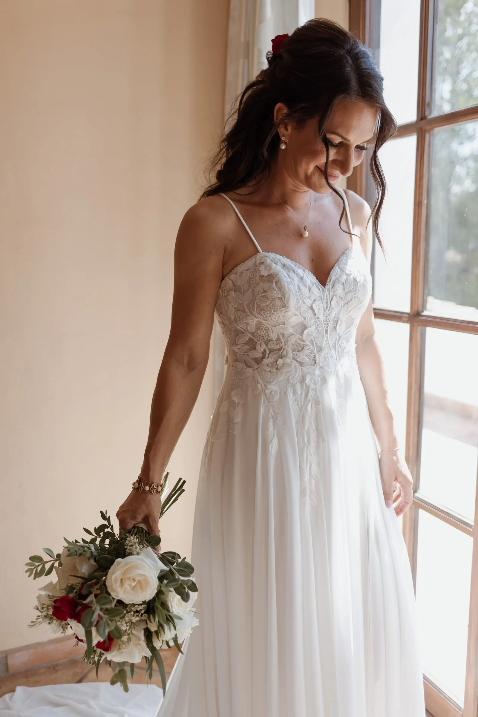 The bride looking down at her dress by a tall window, bouquet in hand, Hacienda San José, Mijas