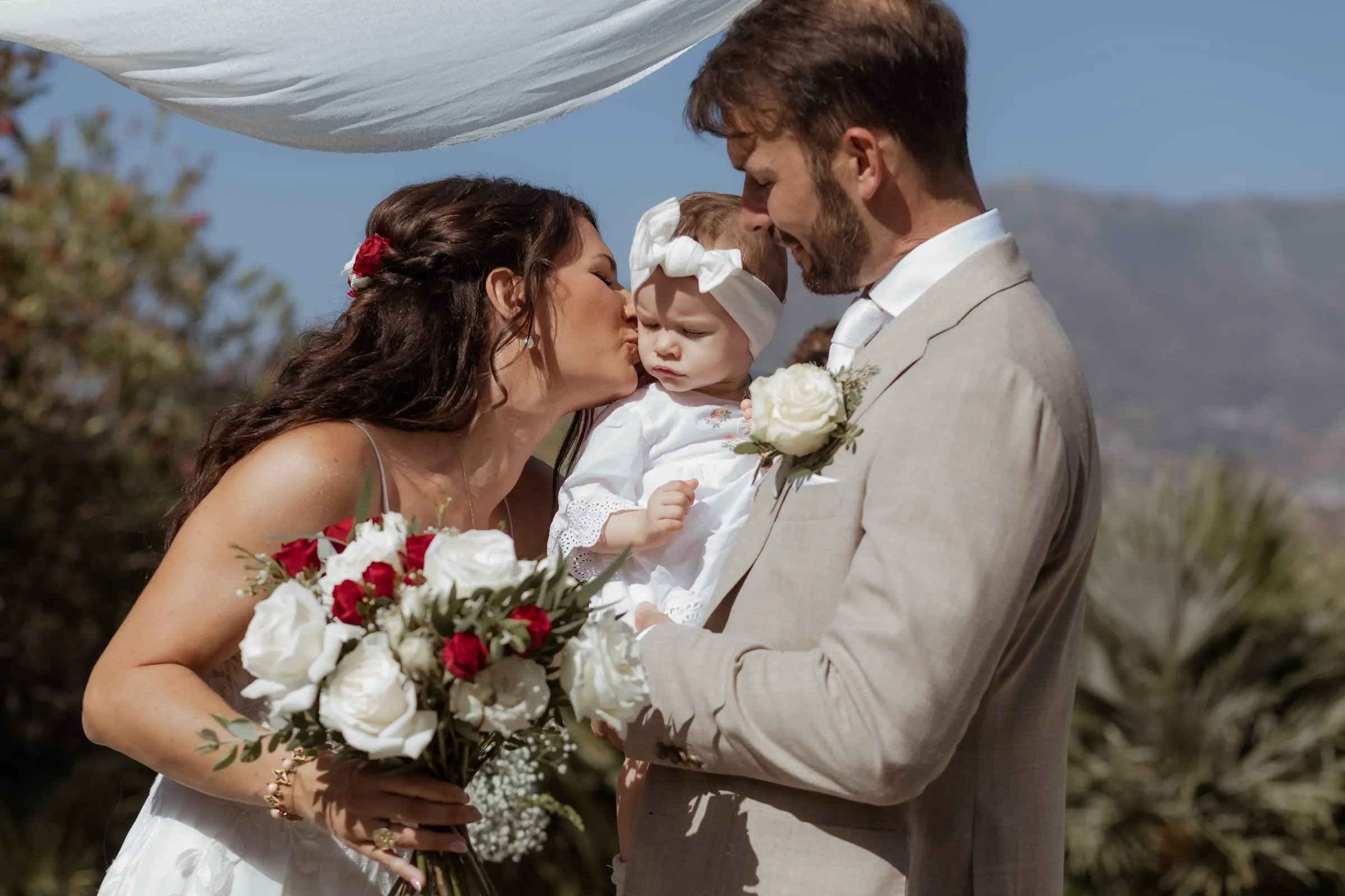 The newlyweds kissing their baby daughter in a white bow under the ceremony canopy, Hacienda San José, Mijas