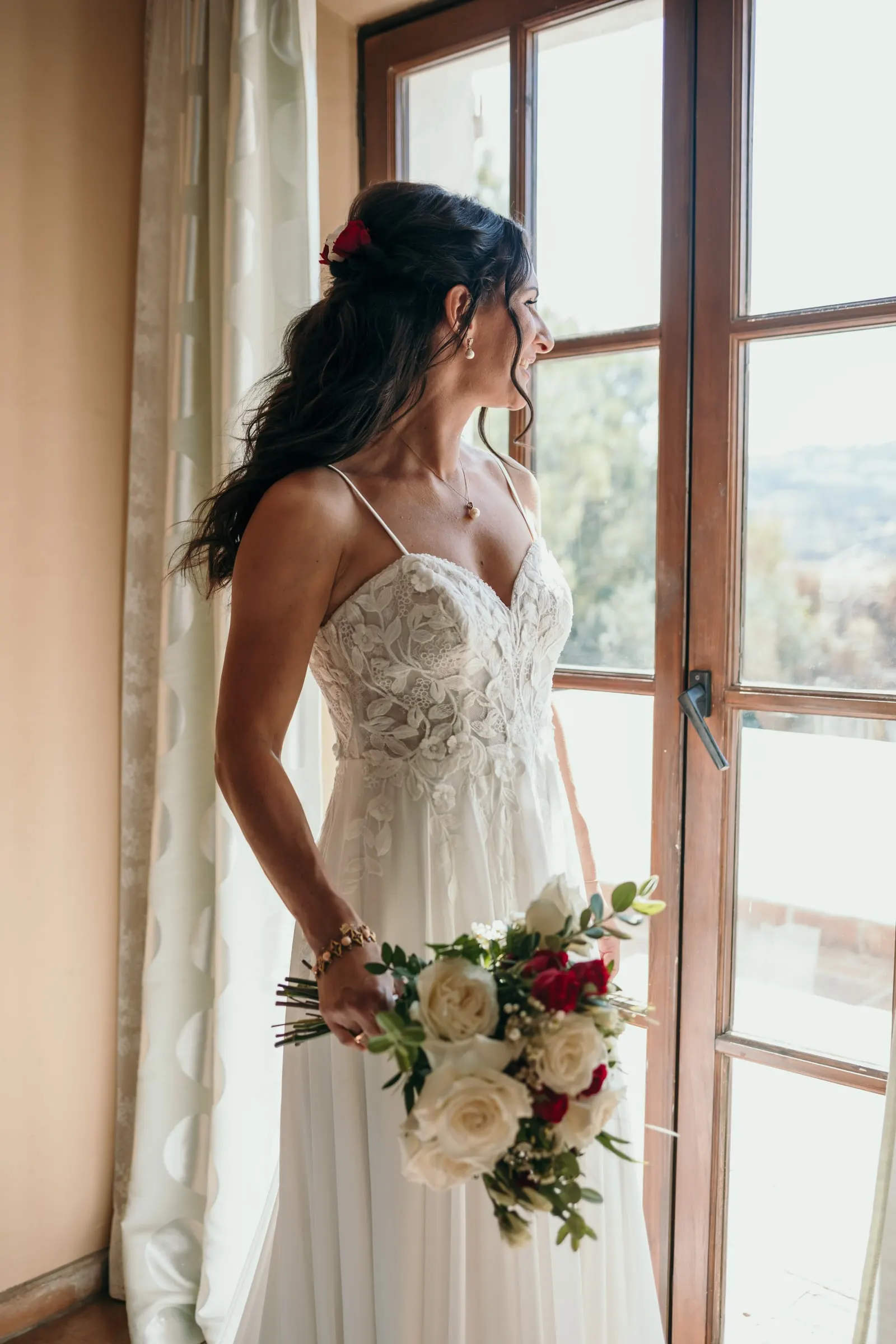 The bride gazing out of a tall window holding her bouquet of roses, Hacienda San José, Mijas