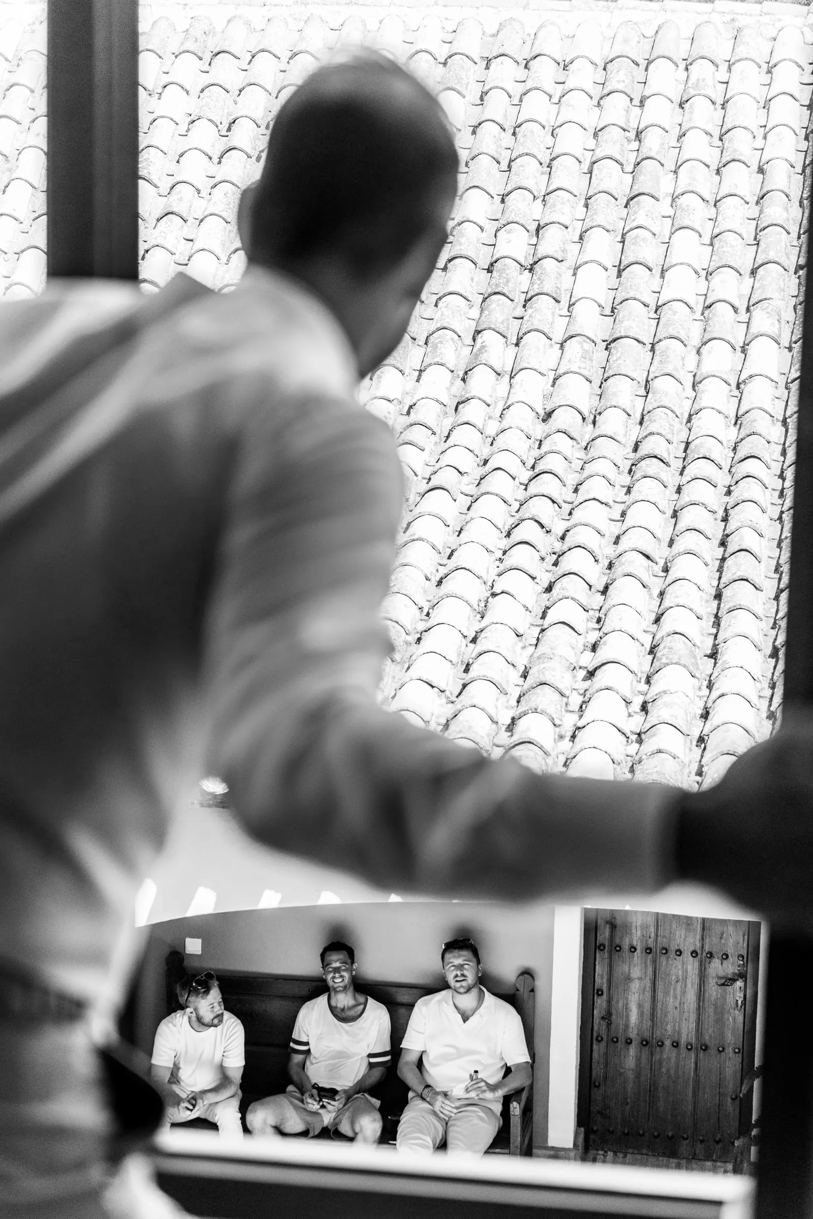 Black and white of the groom looking out over the tiled roofs as friends wait below, Hacienda San José, Mijas