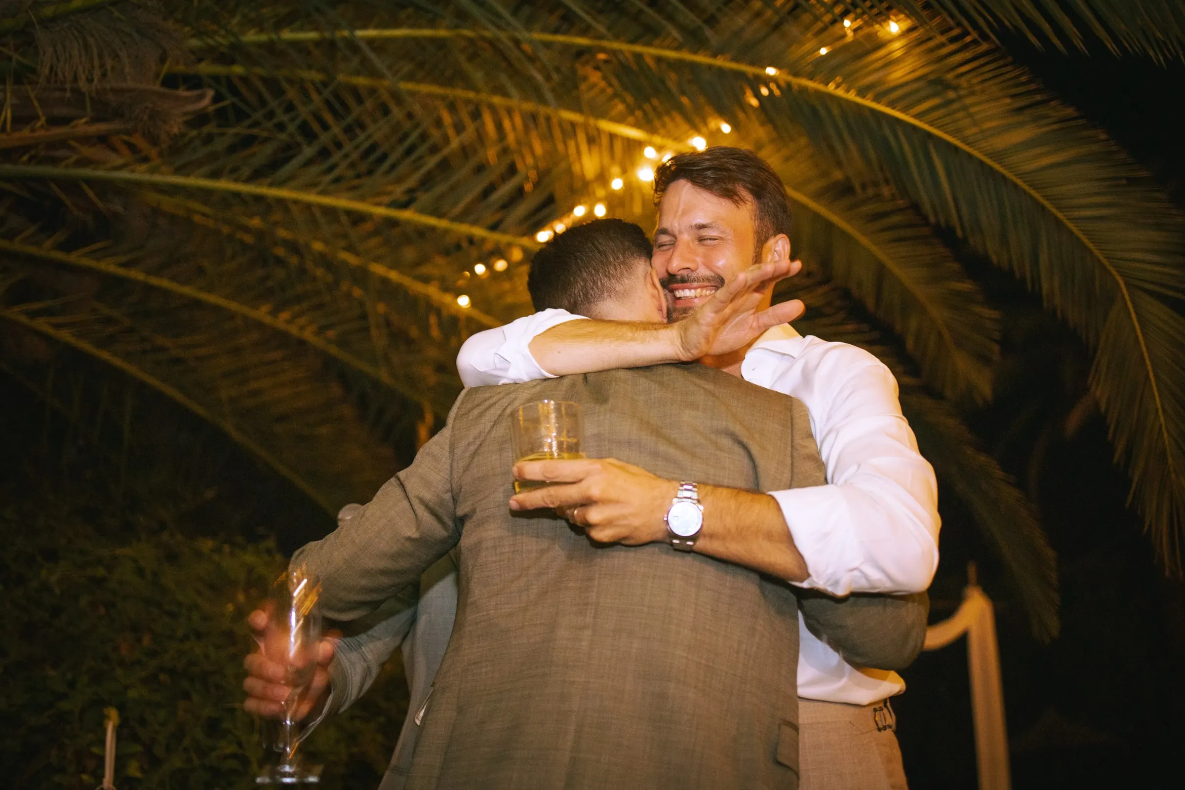 The groom being hugged by a friend under palm fronds and fairy lights, Hacienda San José, Mijas