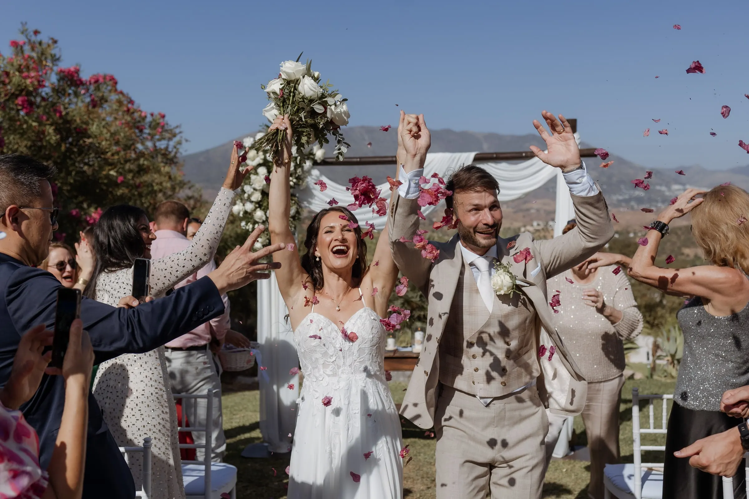 Bride and groom laughing under falling red petals as guests reach out to them, Hacienda San José, Mijas