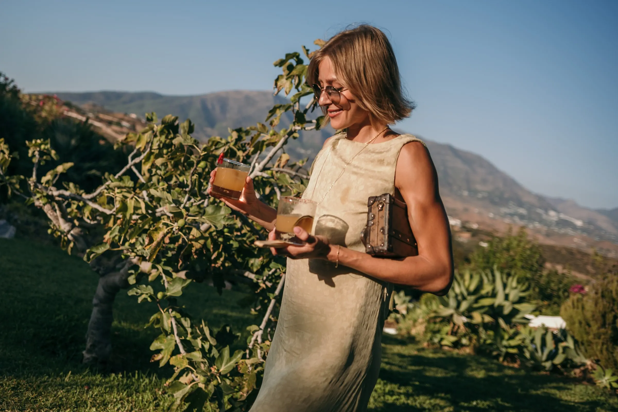 A guest in a linen dress holding drinks beside a fig tree, hills behind, Hacienda San José, Mijas