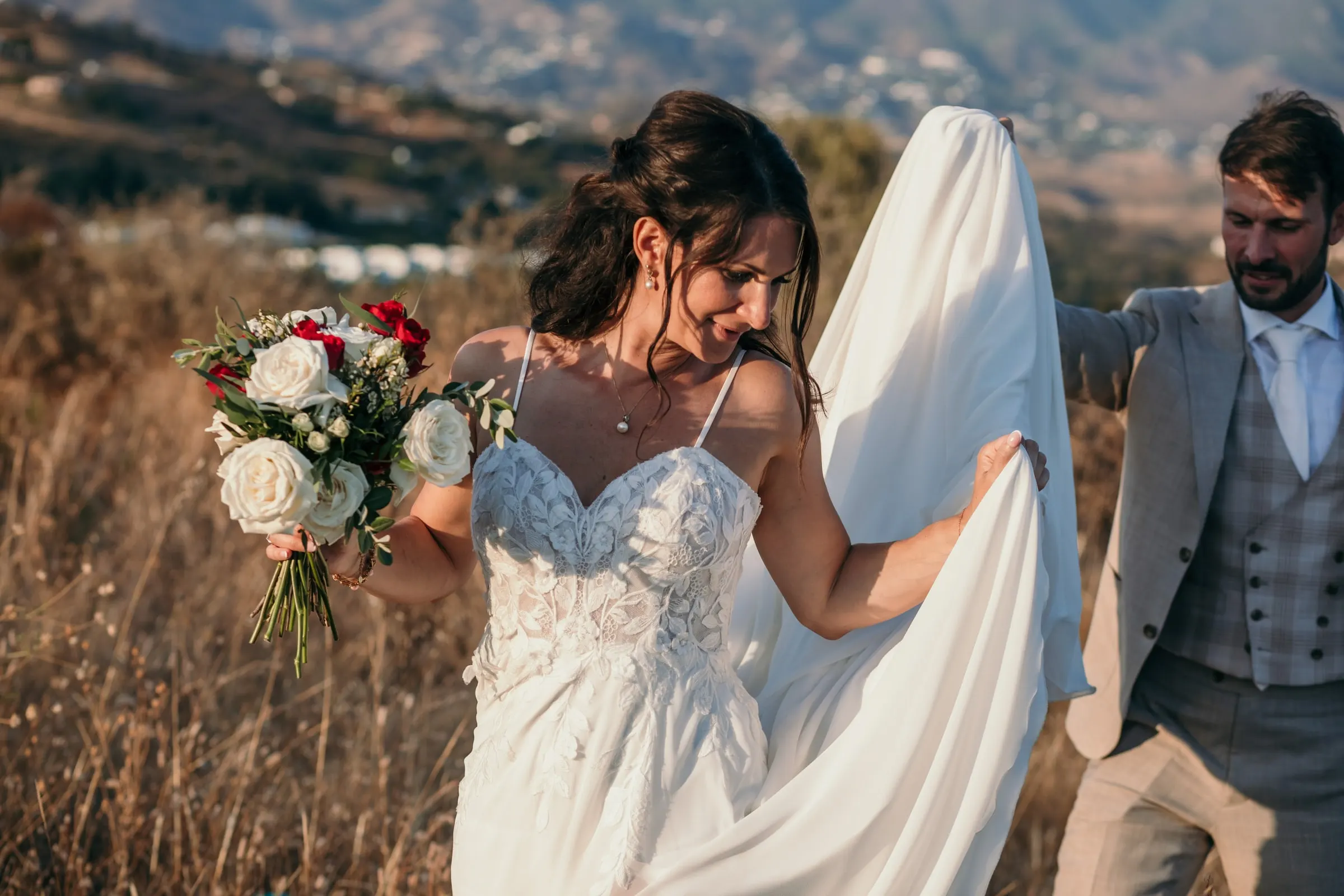 The bride laughing as the groom lifts the train of her dress in a dry field, Hacienda San José, Mijas