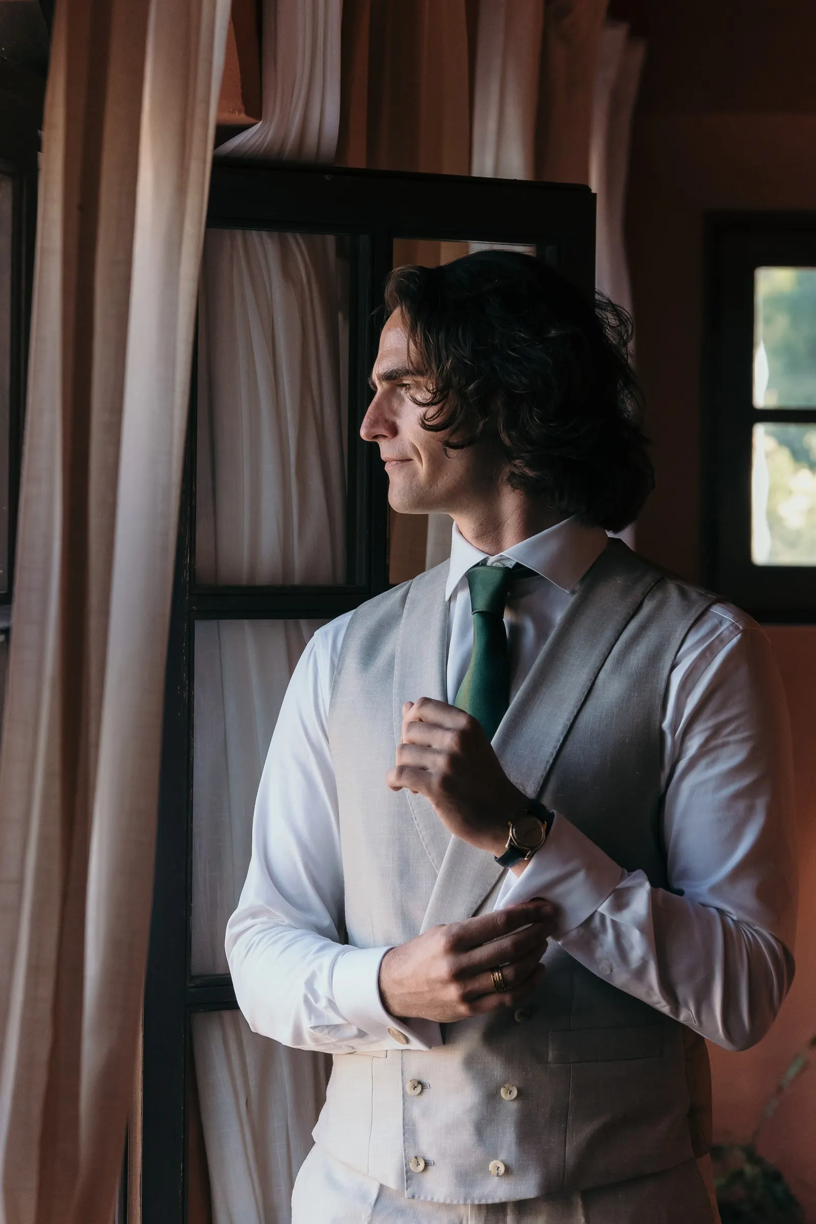 The groom adjusting his cuff by the window in a grey waistcoat and green tie, Hacienda San José, Mijas