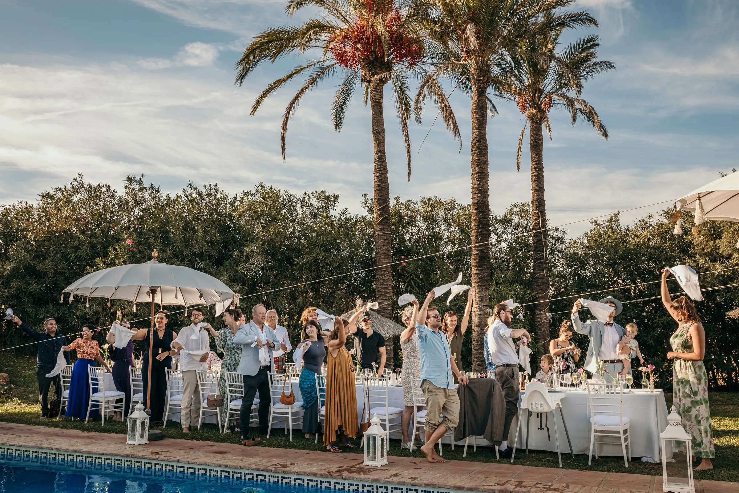 Guests at long tables by the pool waving napkins under tall palm trees, Hacienda San José, Mijas