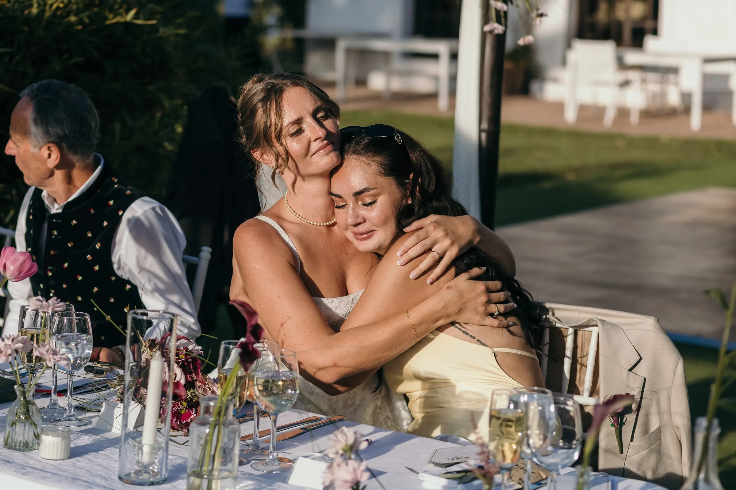 The bride hugging a friend tightly at the dinner table as a guest looks on, Hacienda San José, Mijas