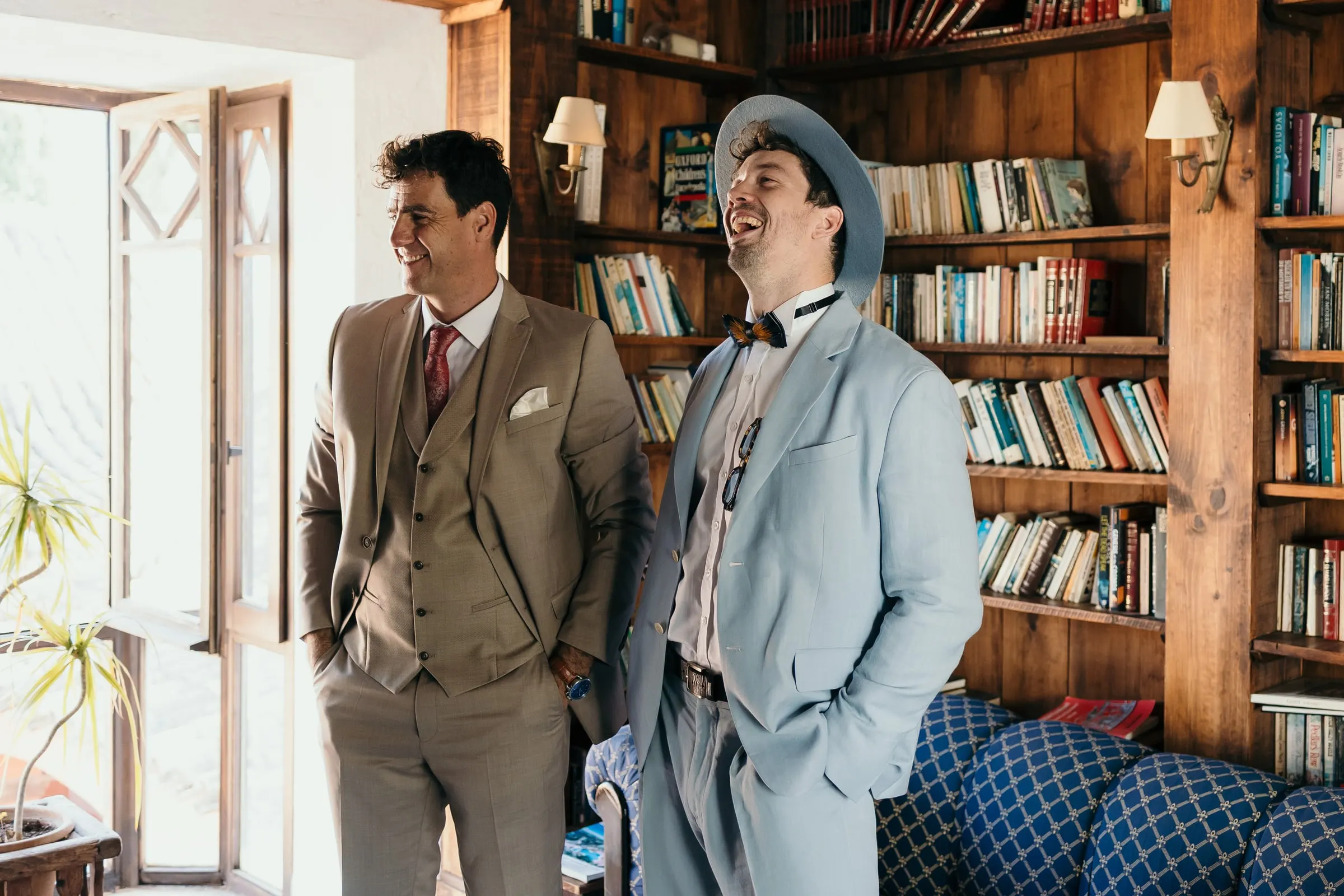 Two groomsmen laughing in the wood-panelled library, one in a pale blue suit, Hacienda San José, Mijas