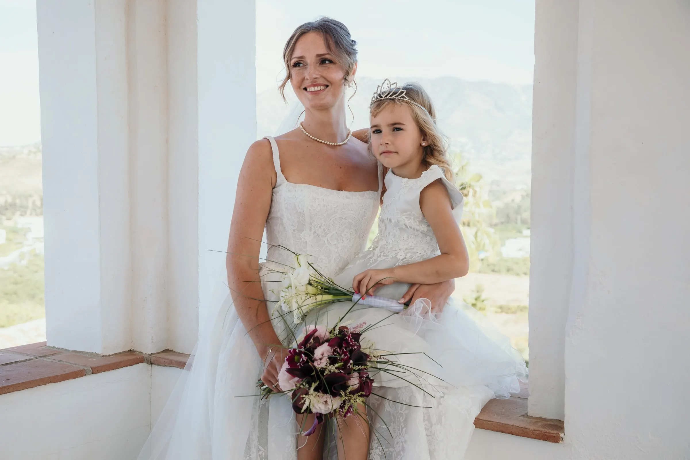The bride sitting on a white window ledge with the flower girl in a tiara on her lap, Hacienda San José, Mijas