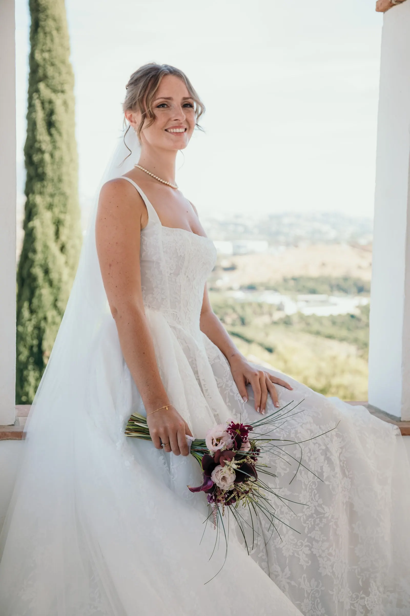 The bride sitting on a whitewashed ledge with a cypress and the valley behind, Hacienda San José, Mijas