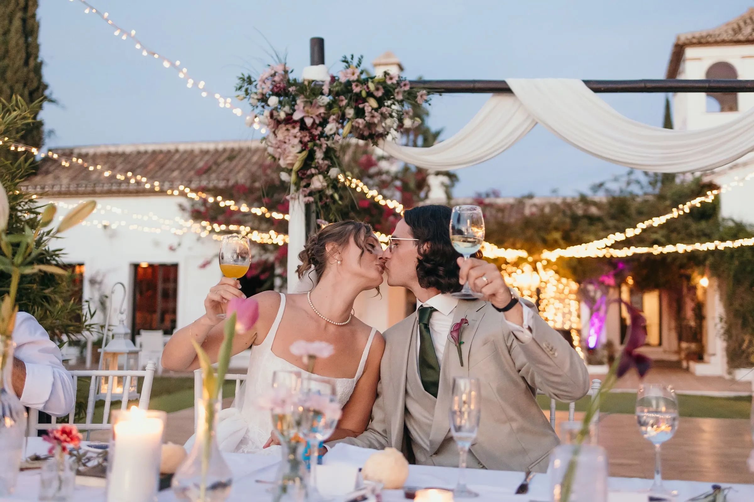 Bride and groom kissing at the head table, glasses in hand, under festoon lights at Hacienda San José, Mijas