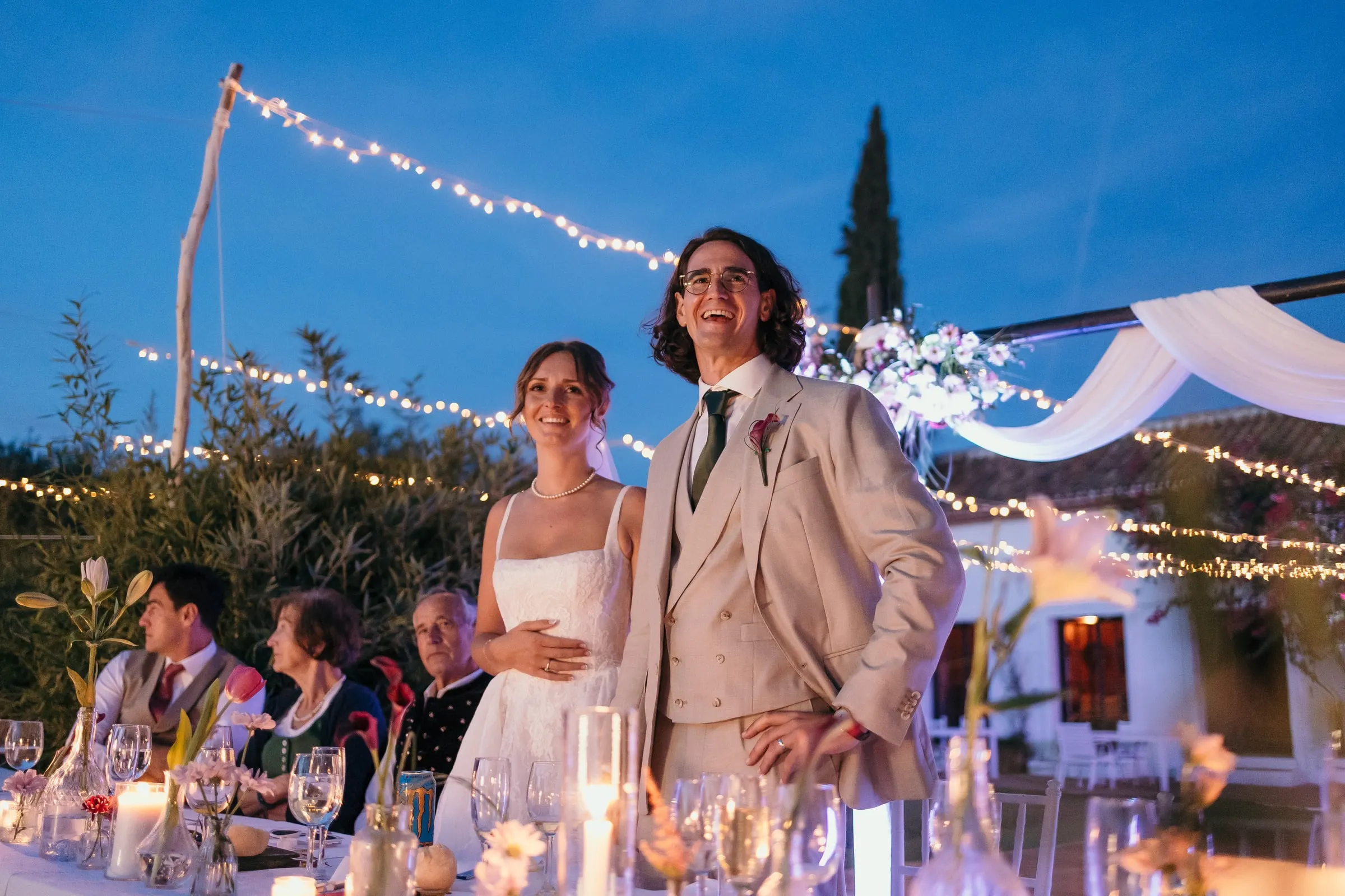Bride and groom standing and laughing at the head table under string lights at dusk at Hacienda San José, Mijas