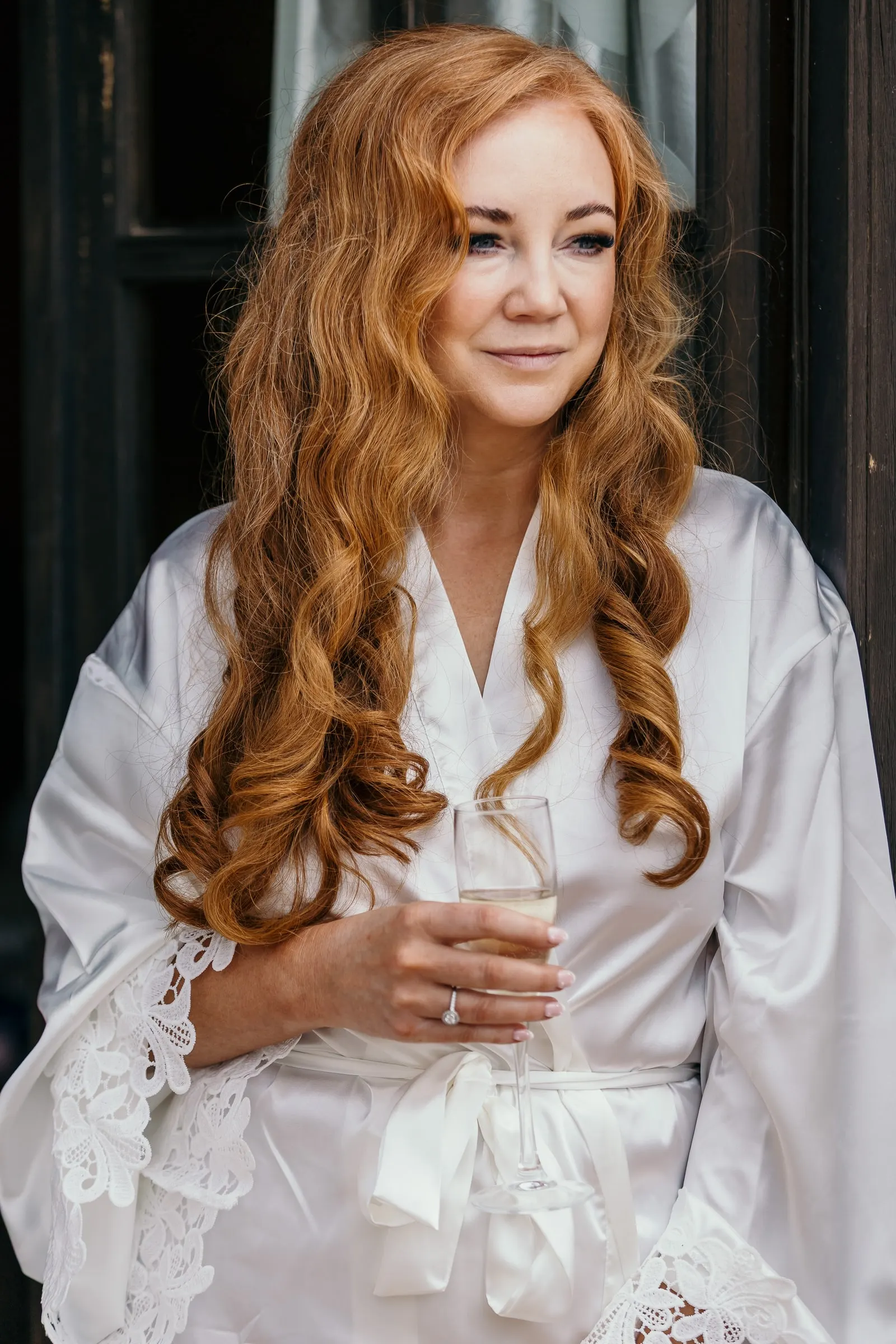 Red-haired bride in a white satin robe holding a glass of cava, wedding morning at Hacienda San José