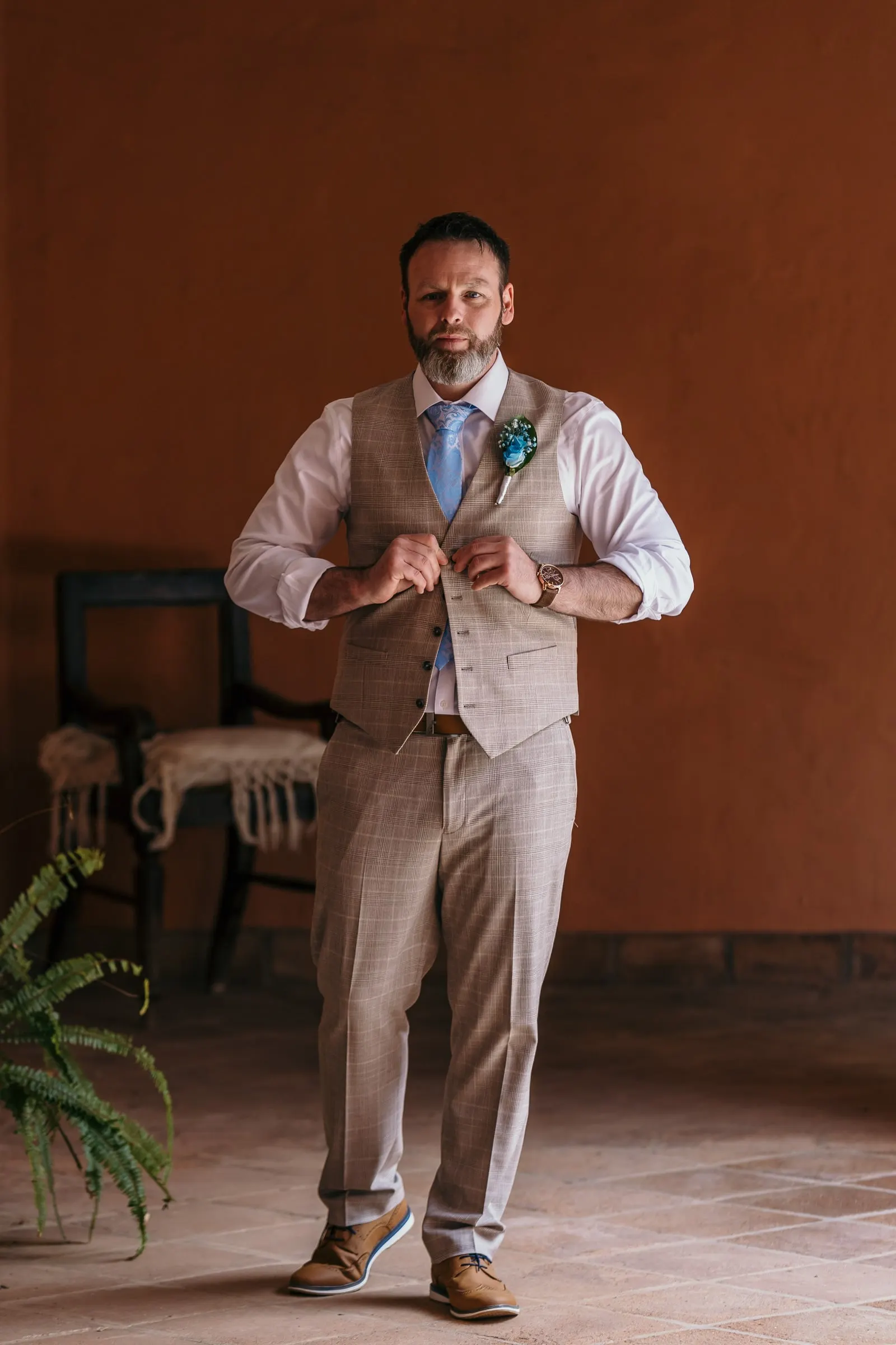 Groom in a beige check waistcoat buttoning his cuffs in a terracotta-walled room at Hacienda San José