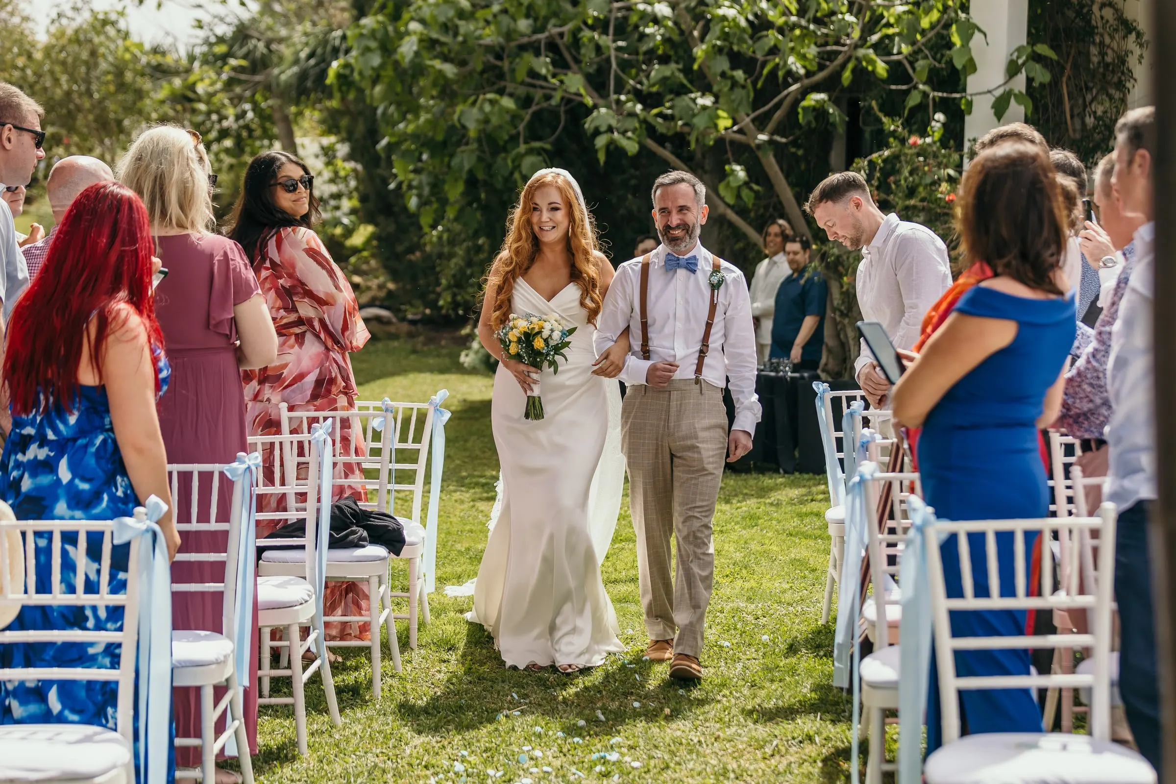 Bride walking down the grass aisle on the arm of a friend in braces, outdoor ceremony in Mijas