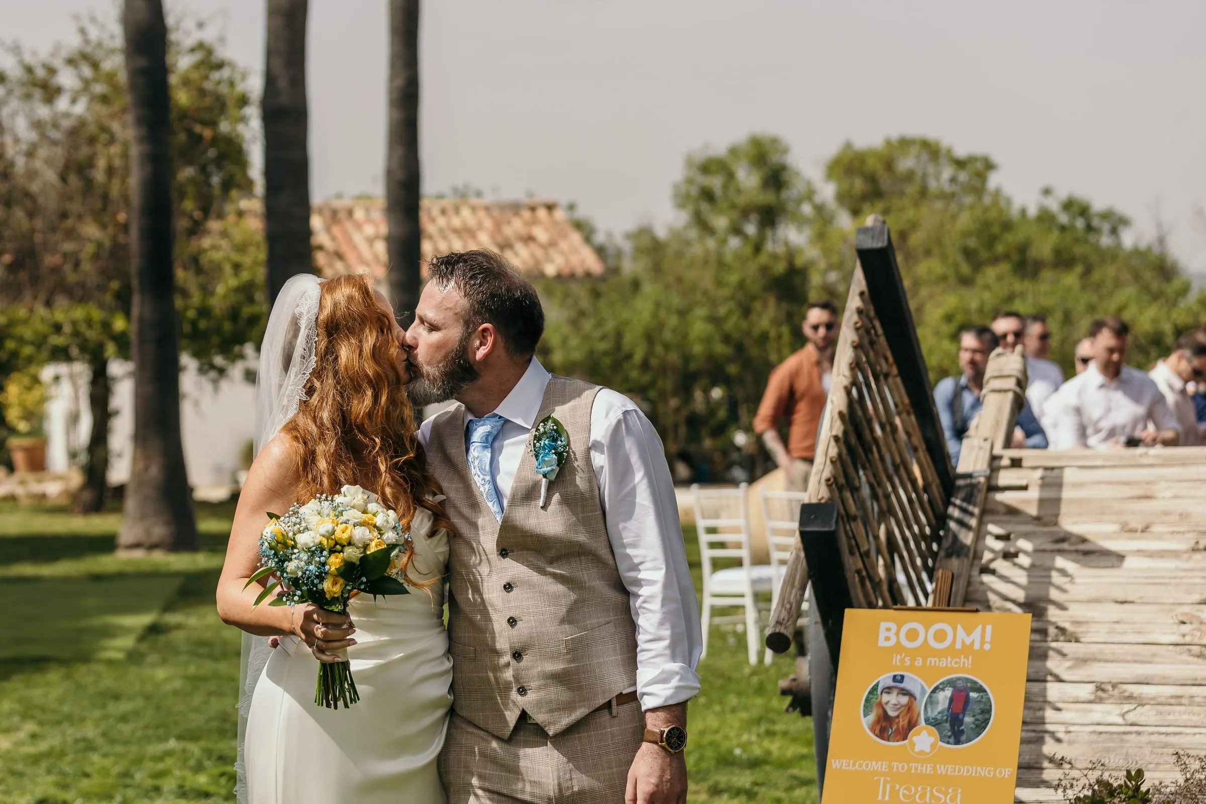 Groom kissing the bride on the cheek beside a wooden sign on the lawn, Mijas