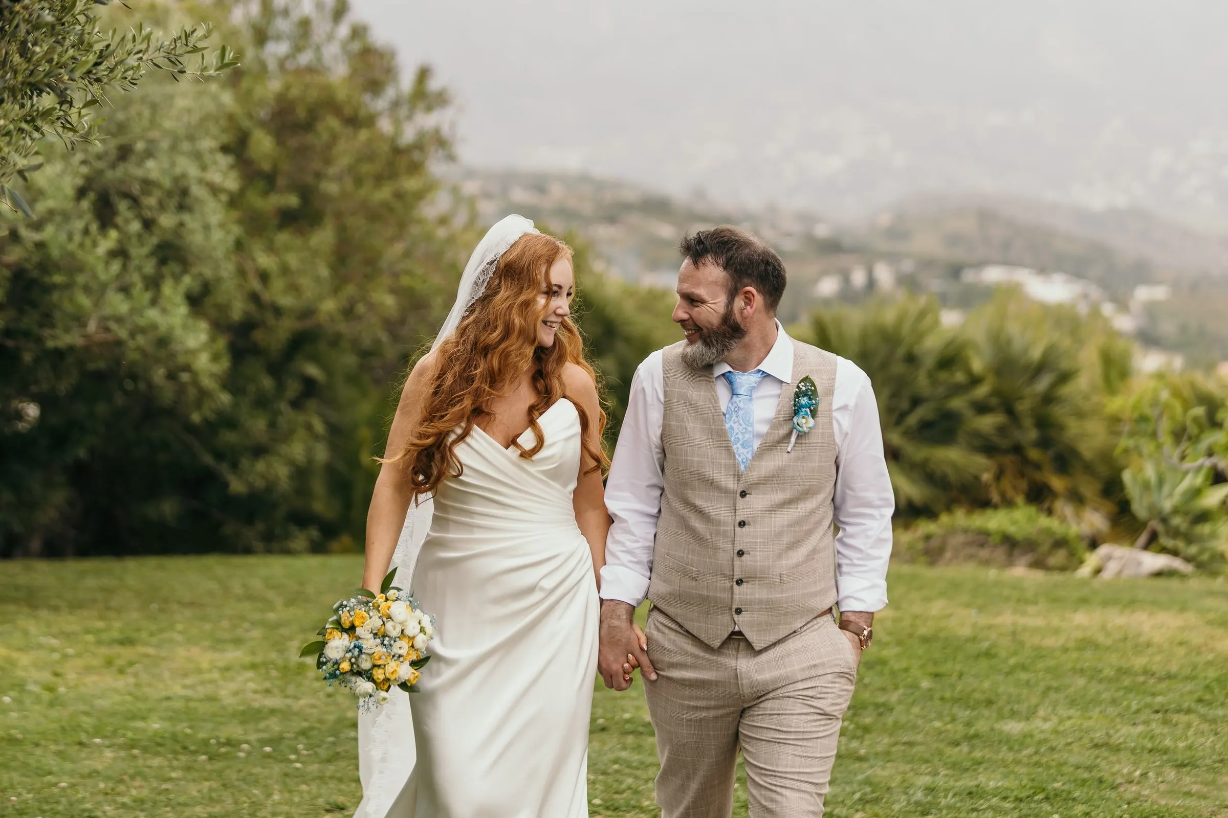 Bride and groom laughing as they walk across the garden with the hills of Mijas behind