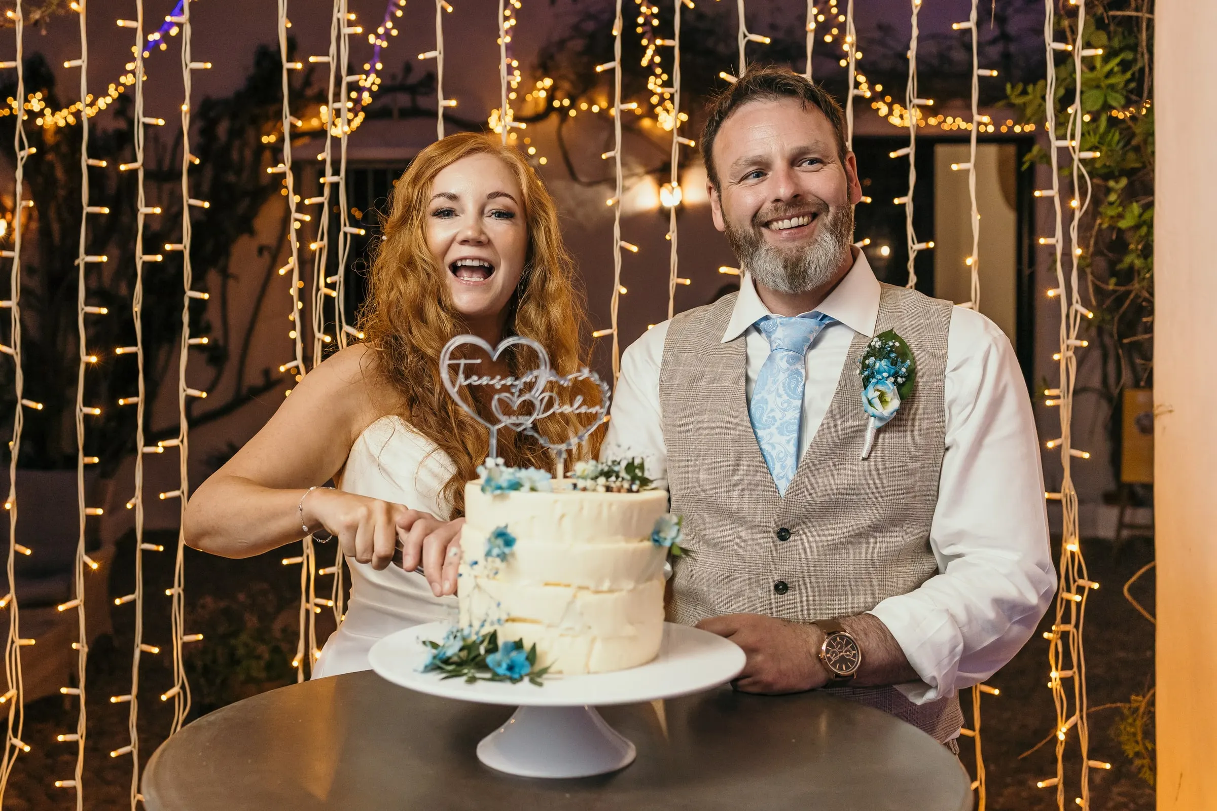 Couple cutting a white wedding cake with blue flowers under a curtain of fairy lights, Hacienda San José