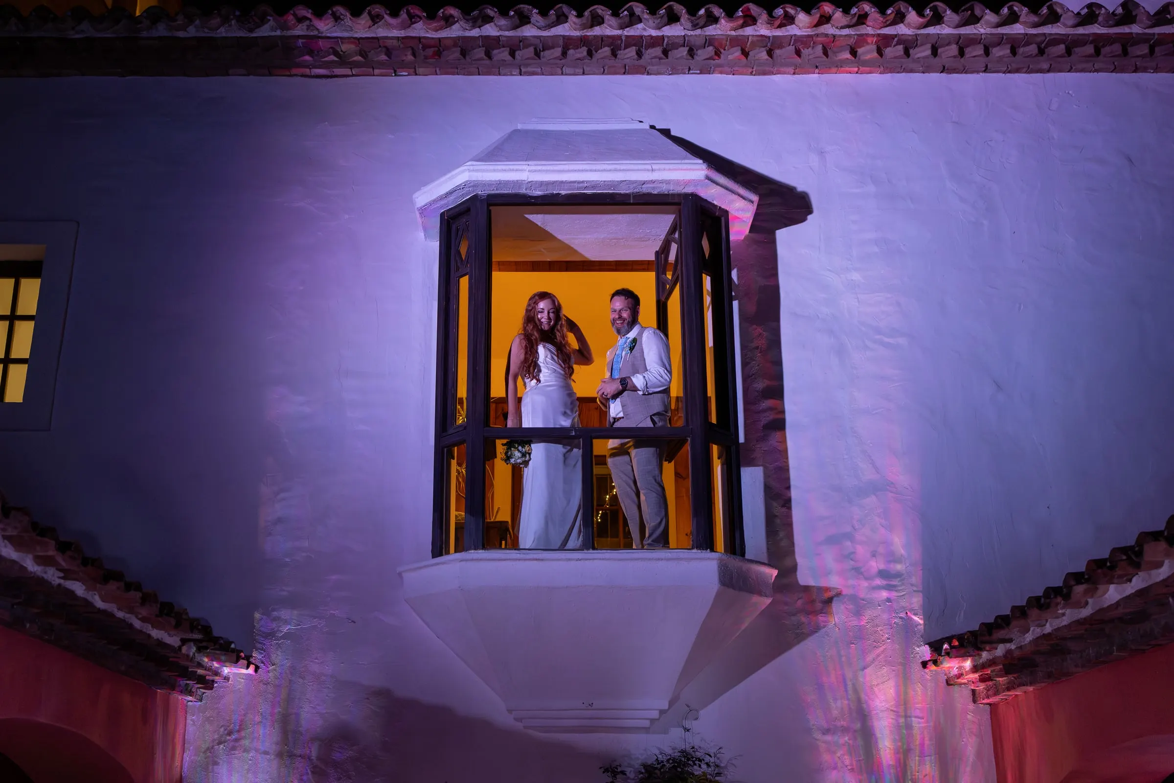 Bride and groom in a lit window bay on the purple-lit white façade of Hacienda San José