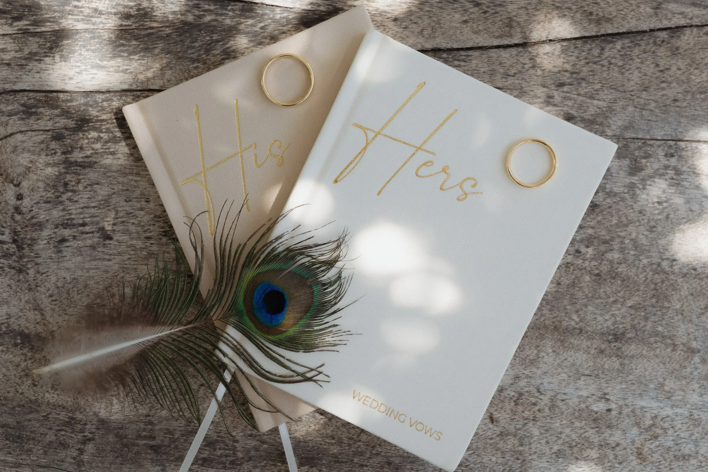His and Hers vow books with a peacock feather on weathered wood