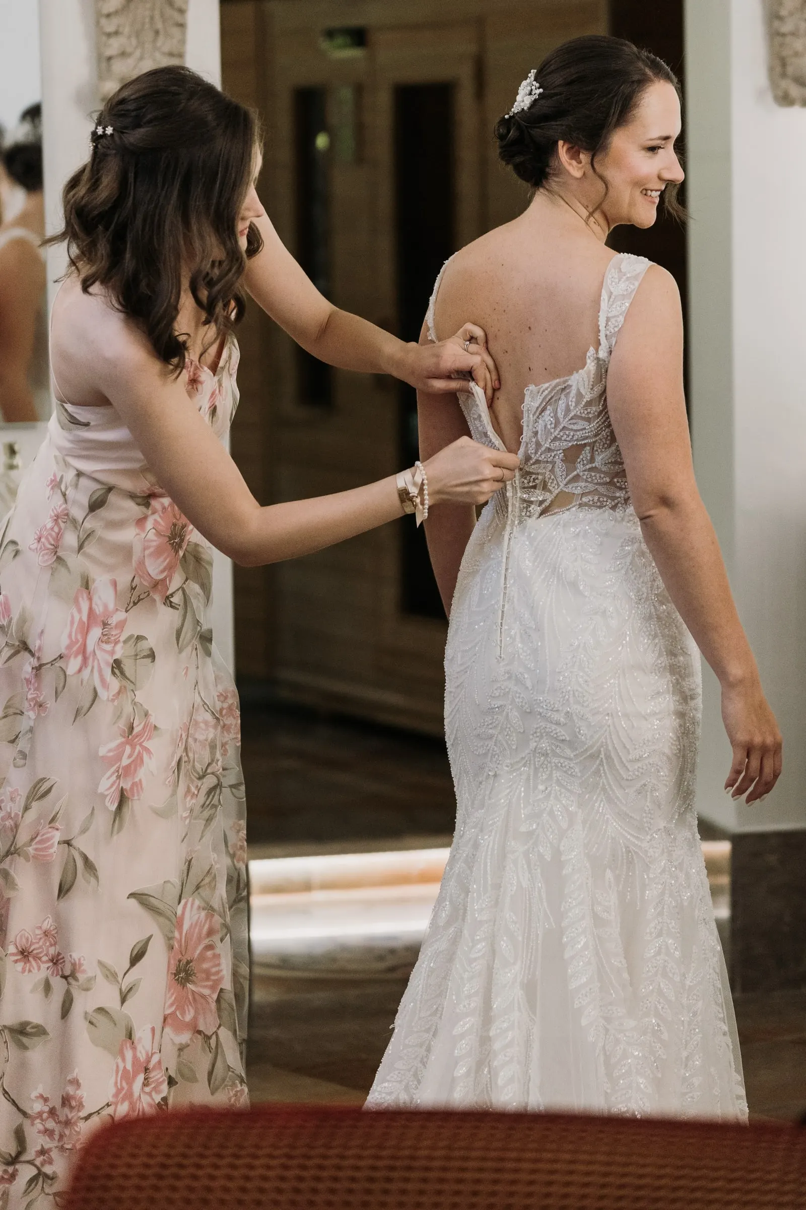 Bridesmaid in a floral dress buttoning the back of the bride's lace gown