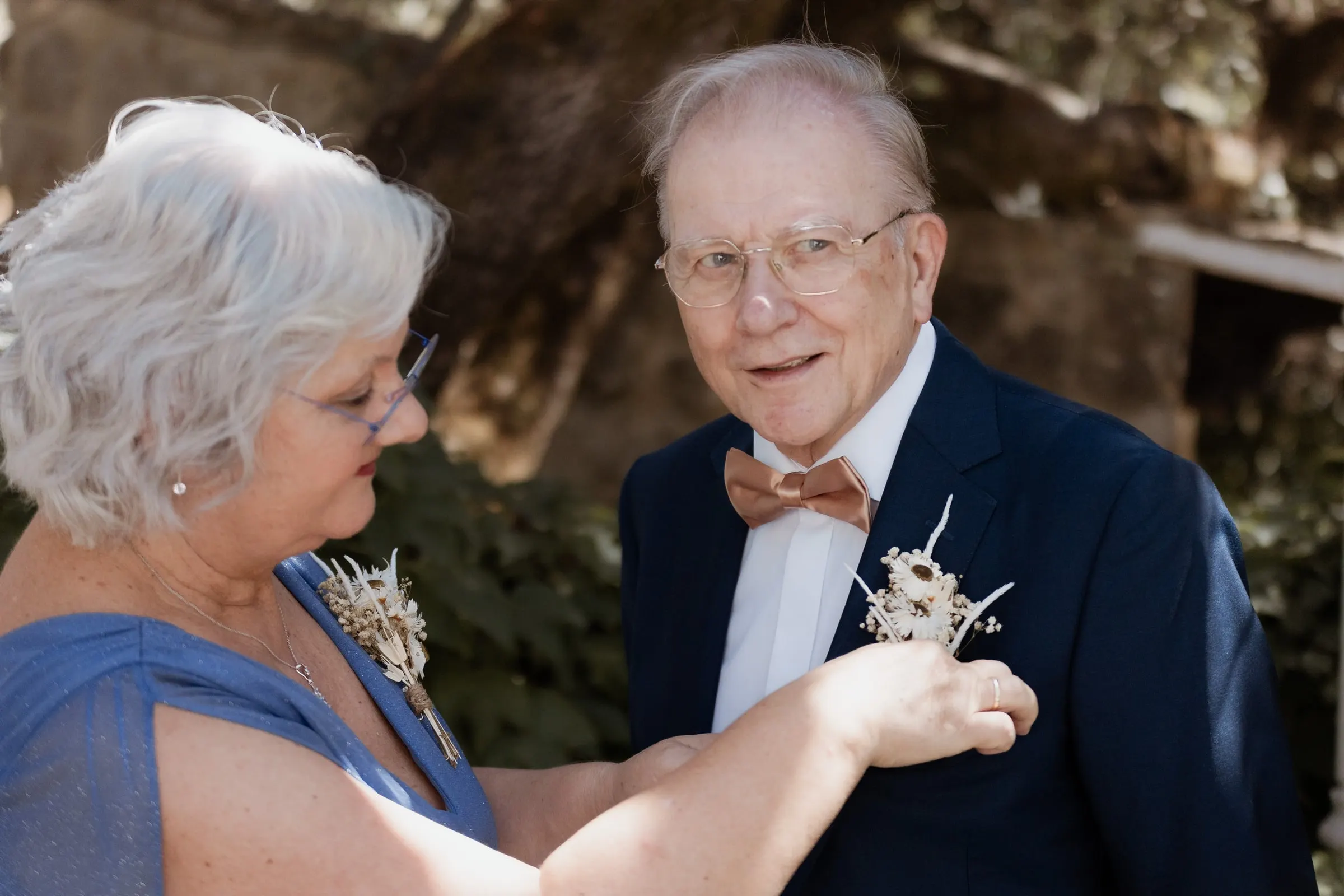 Older woman pinning a dried-flower buttonhole on an older man in a blue suit