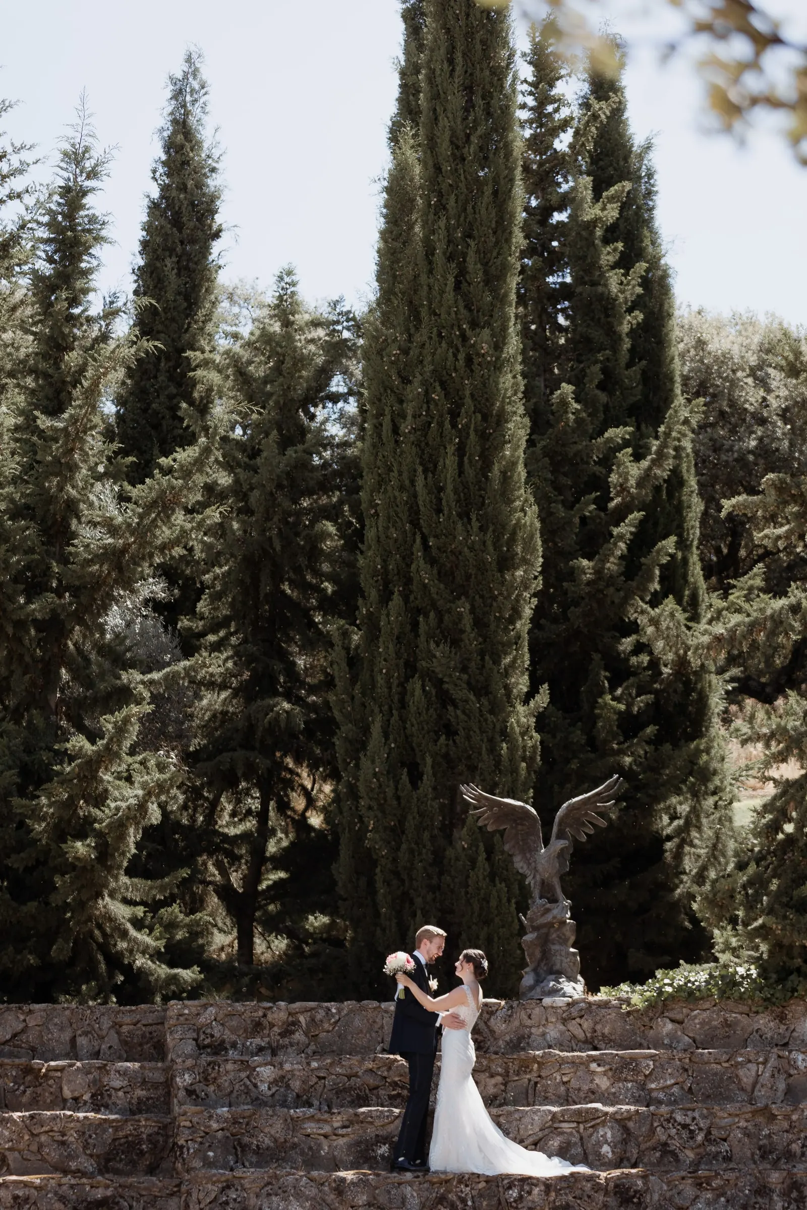 Bride and groom small beneath tall cypress trees and the eagle statue in Ronda
