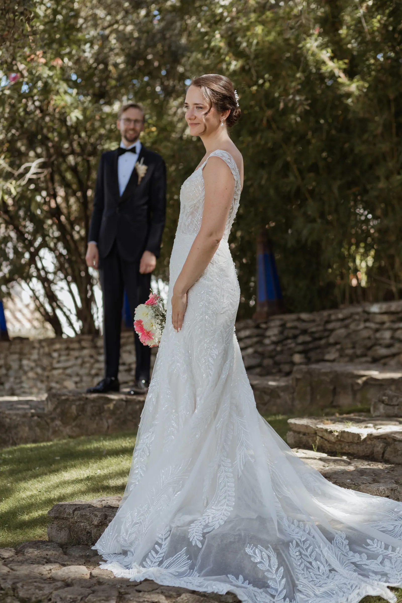 Bride with her long train on the stone terrace, groom waiting behind her