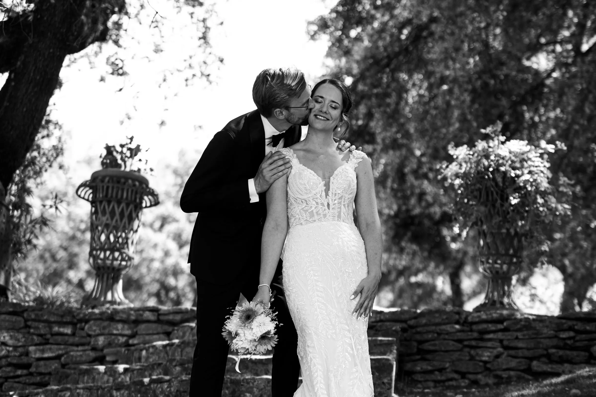 Black and white photo of the groom kissing the bride's cheek in the garden