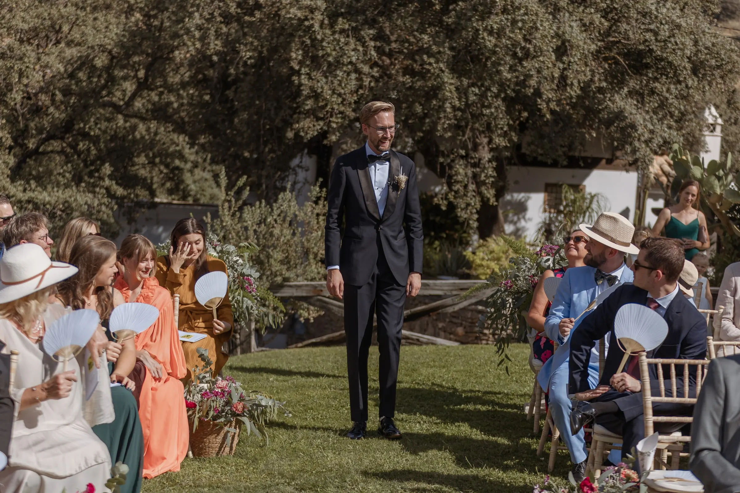 Groom waiting at the end of the aisle as guests cool off with fans