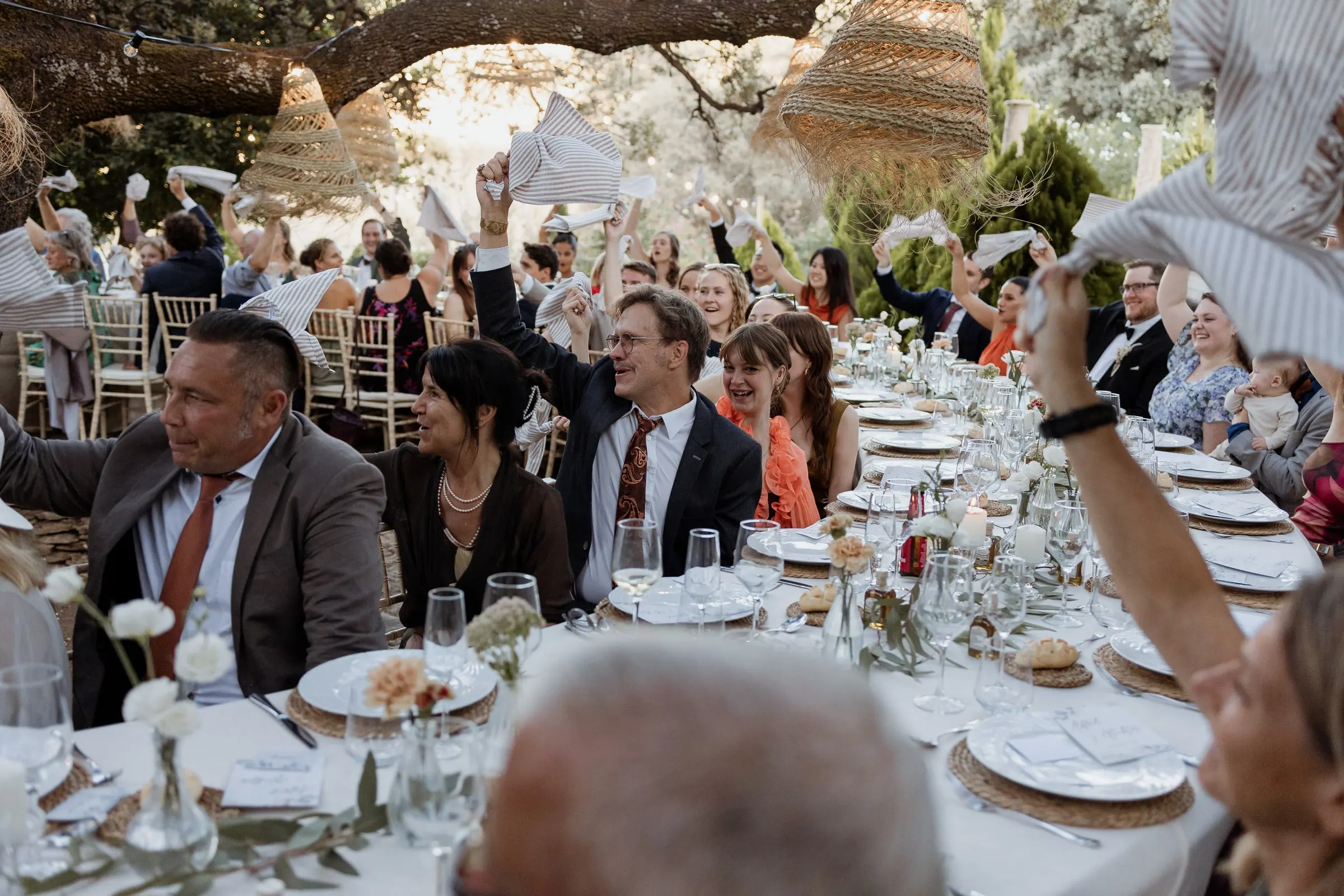 Guests at the long dinner table waving napkins under woven lamps