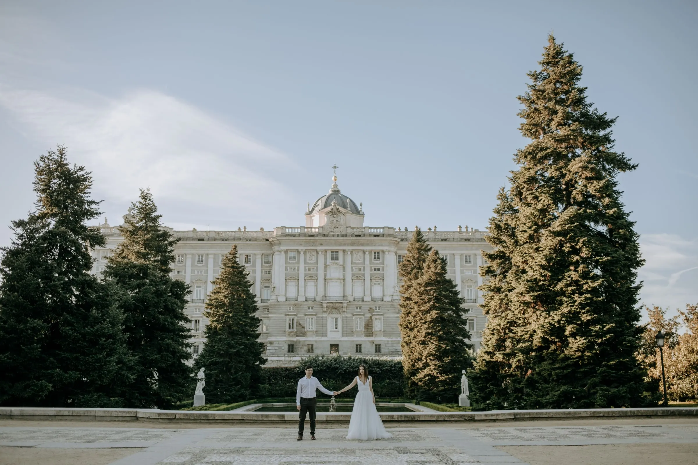 Bride and groom holding hands, tiny in front of the Royal Palace of Madrid
