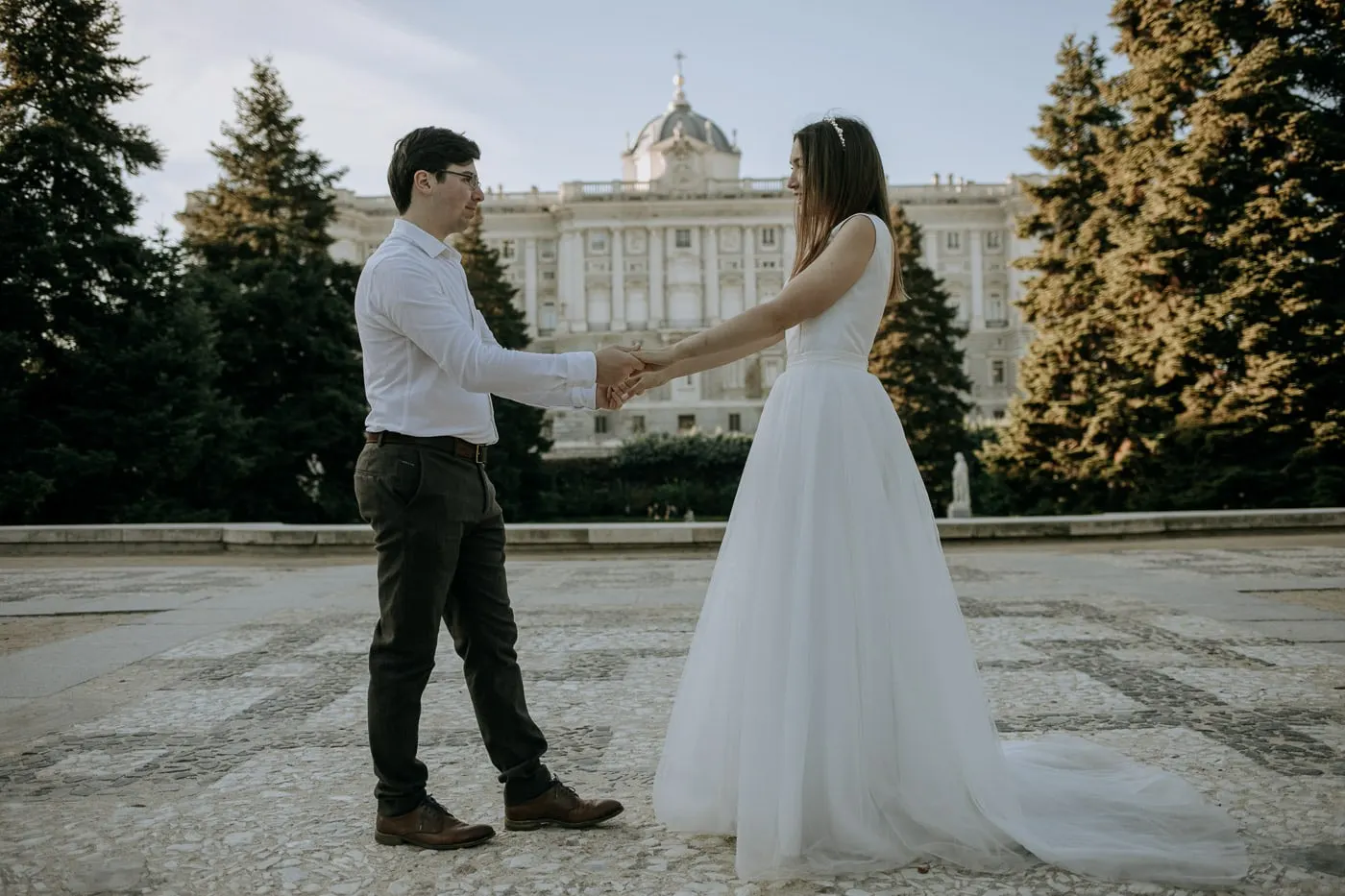 Bride and groom holding hands among fir trees facing the Royal Palace