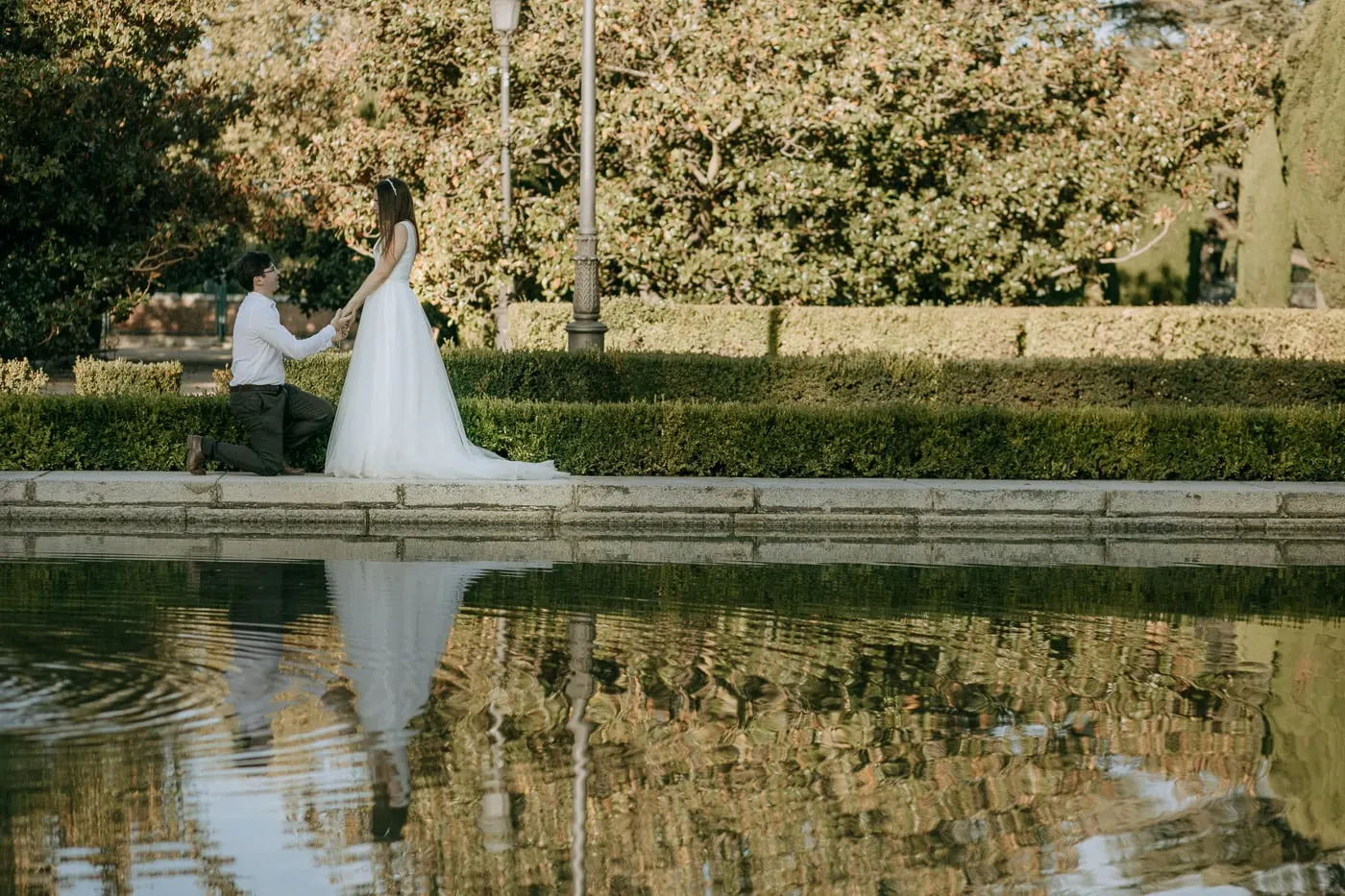 Groom kneeling before the bride at the edge of a garden pond