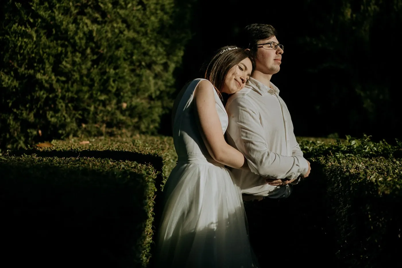 Bride resting her head on the groom's shoulder in the shade of the hedges