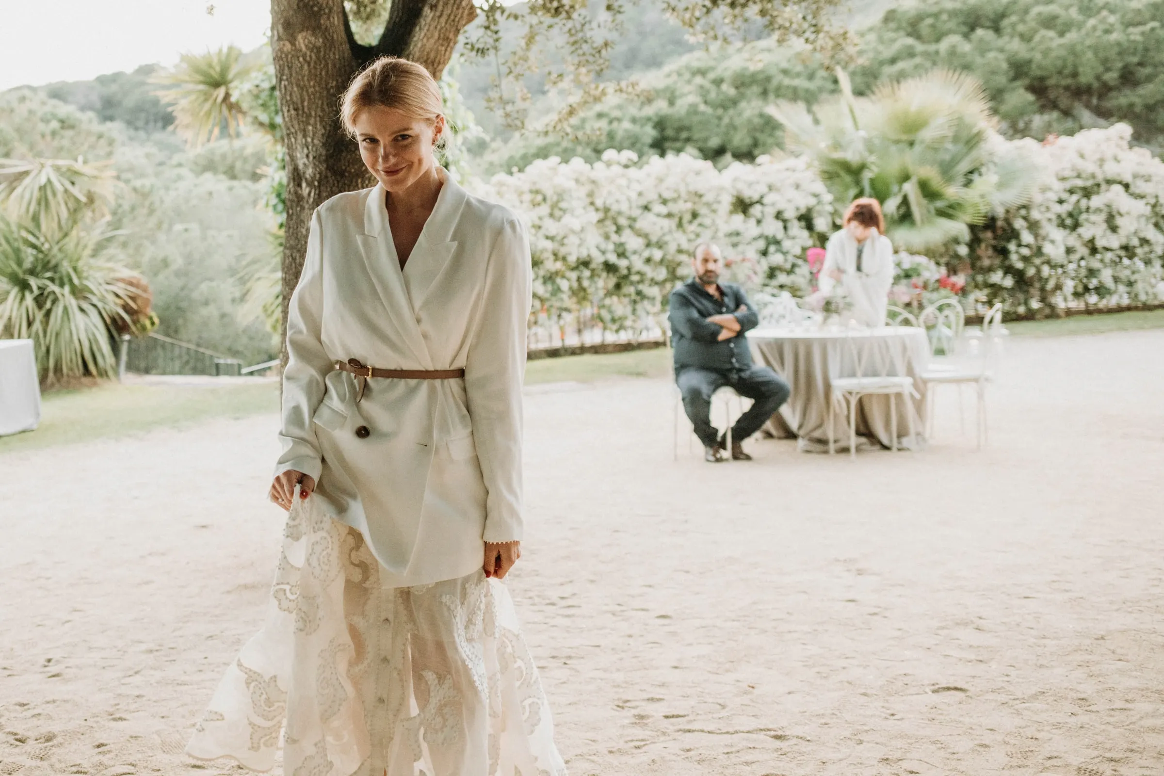 The bride in a white blazer and embroidered skirt smiling in the gravel garden of a masia in Girona