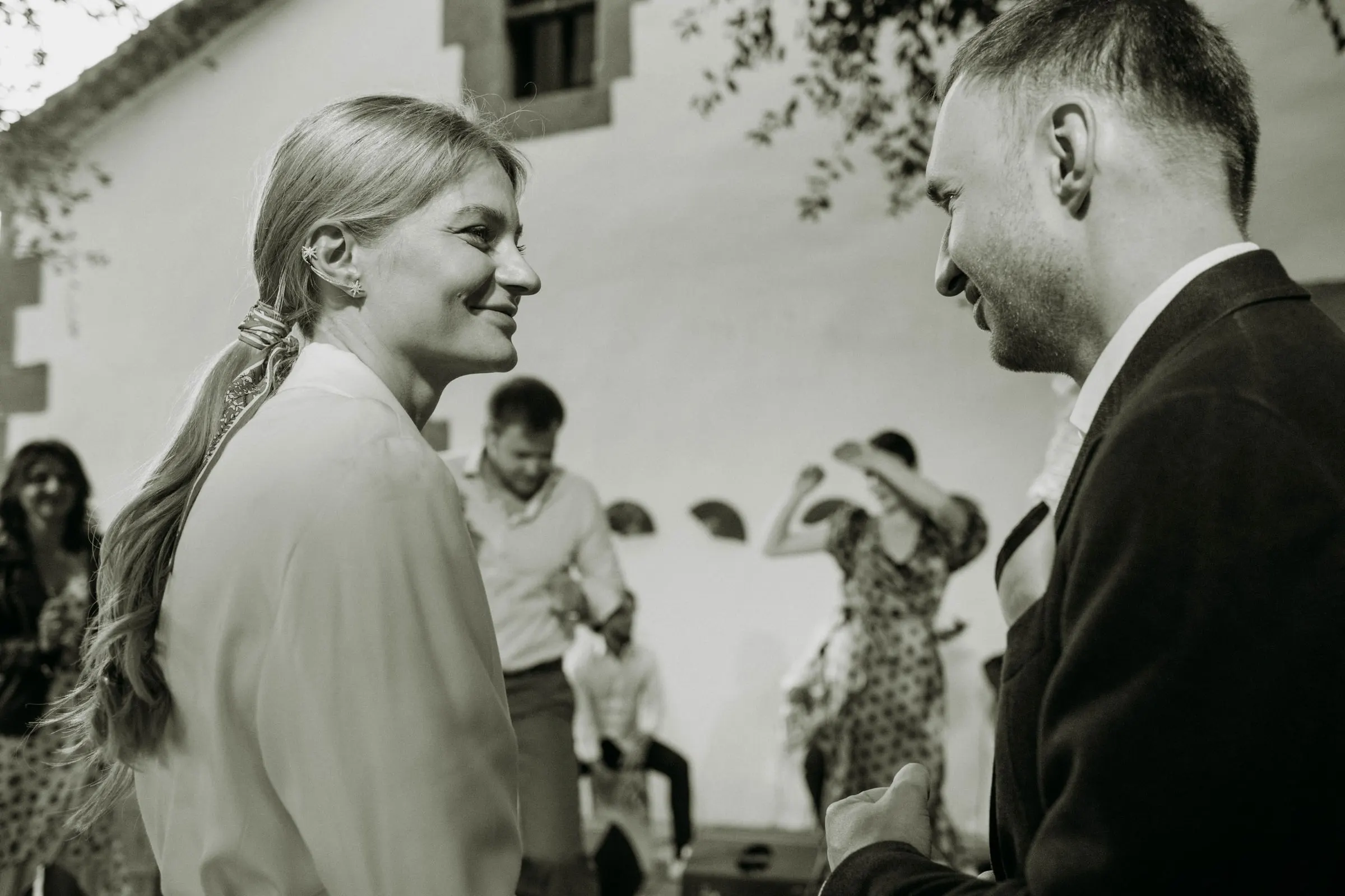 Black and white of the couple smiling at each other as guests dance behind, at a masia in Girona