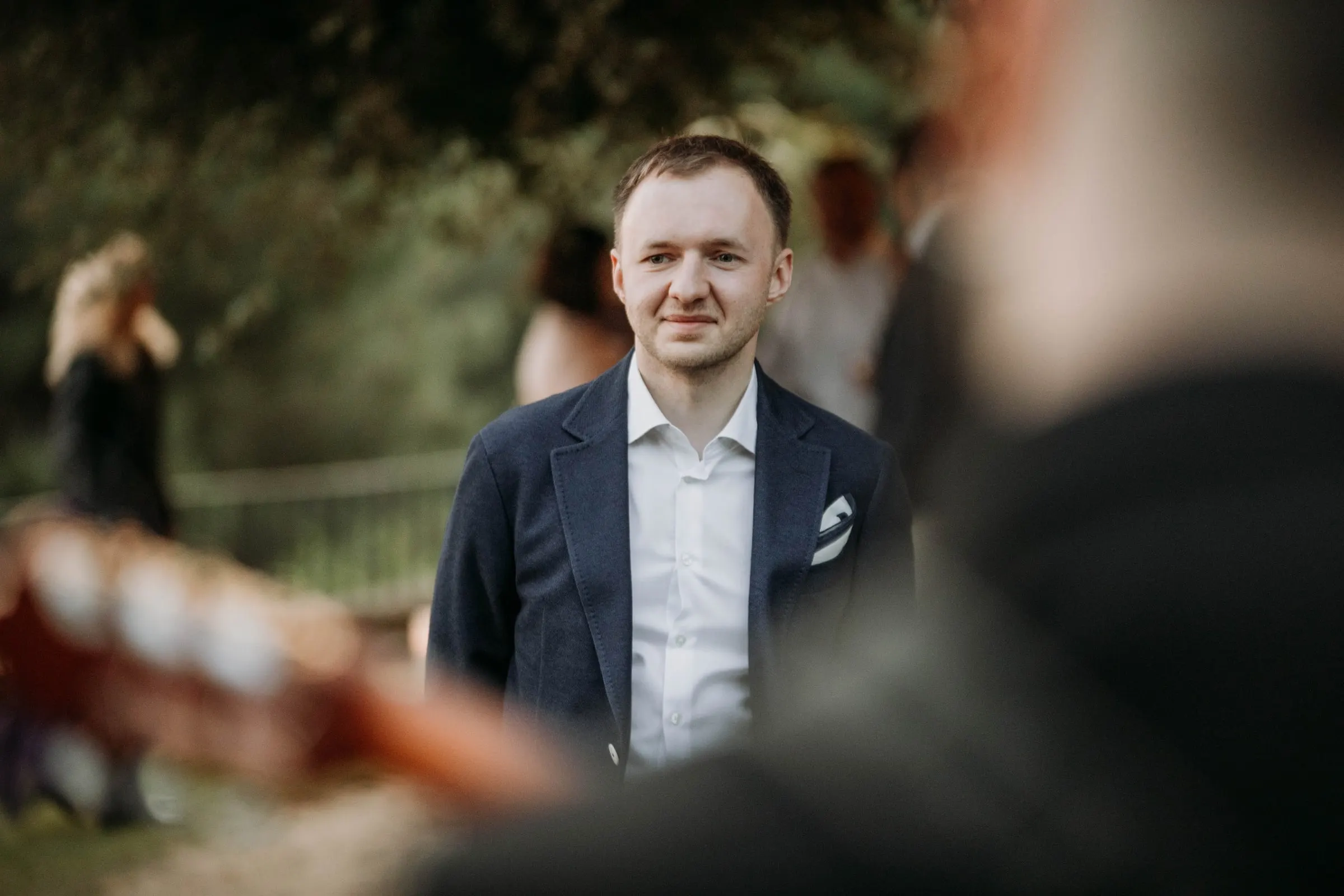 The groom in a navy jacket listening to a guitarist, out of focus in front, at a masia in Girona