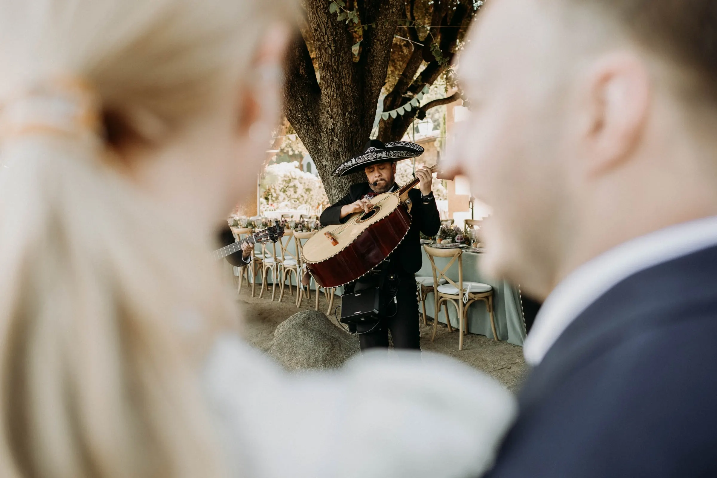 A mariachi playing the guitarrón under a tree, seen between the bride and groom, at a masia in Girona