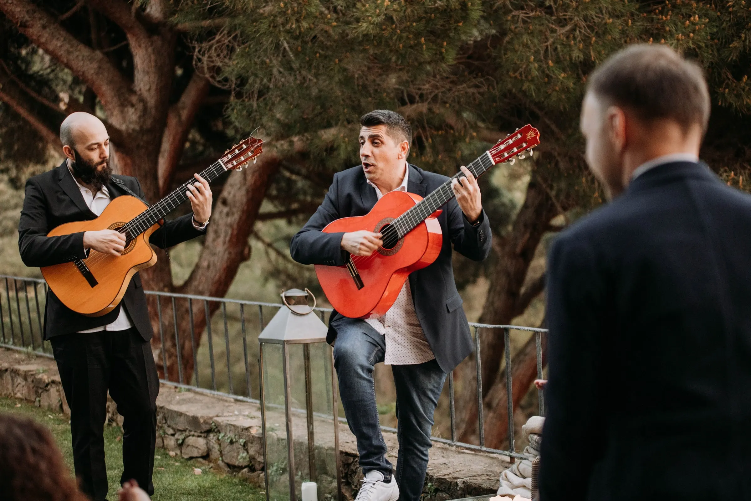 Two guitarists playing in the garden as the groom watches from behind, at a masia in Girona