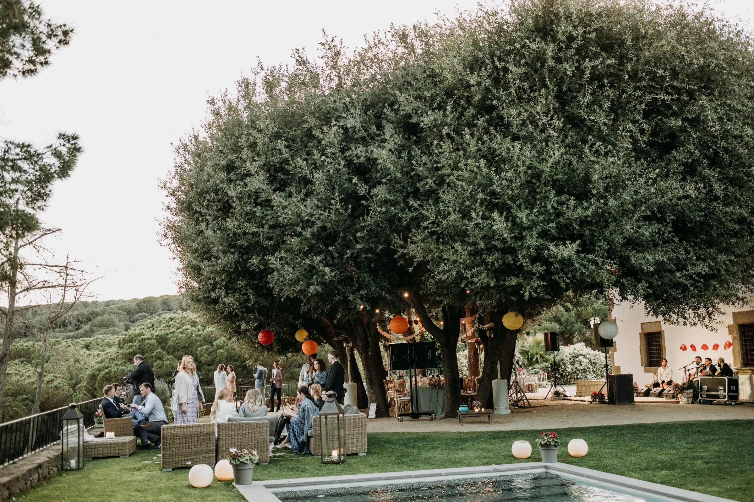 Guests on garden sofas beside the pool under a big tree hung with lanterns, at a masia in Girona