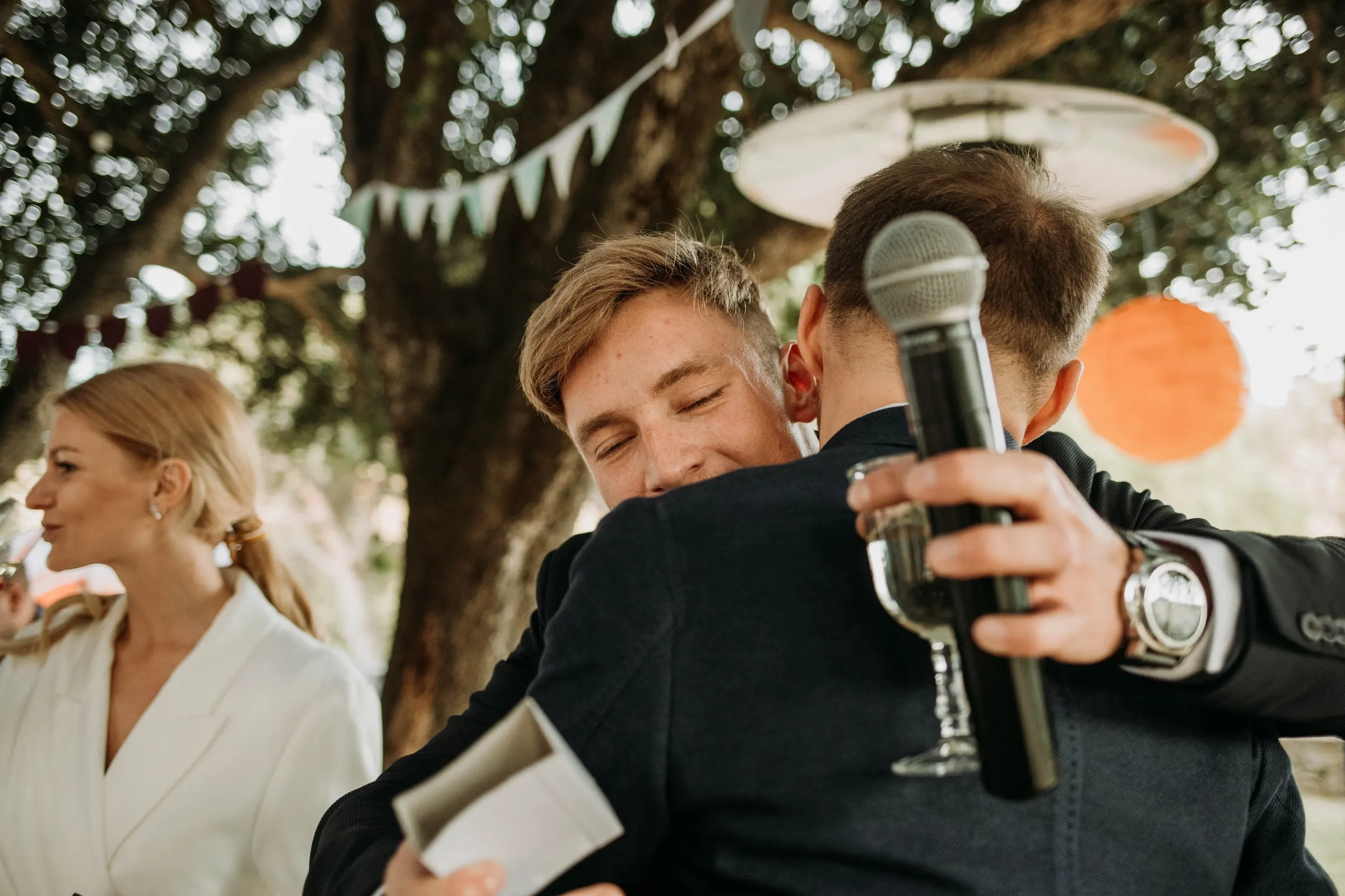 The groom hugging a guest holding a microphone and a glass during the speeches, at a masia in Girona