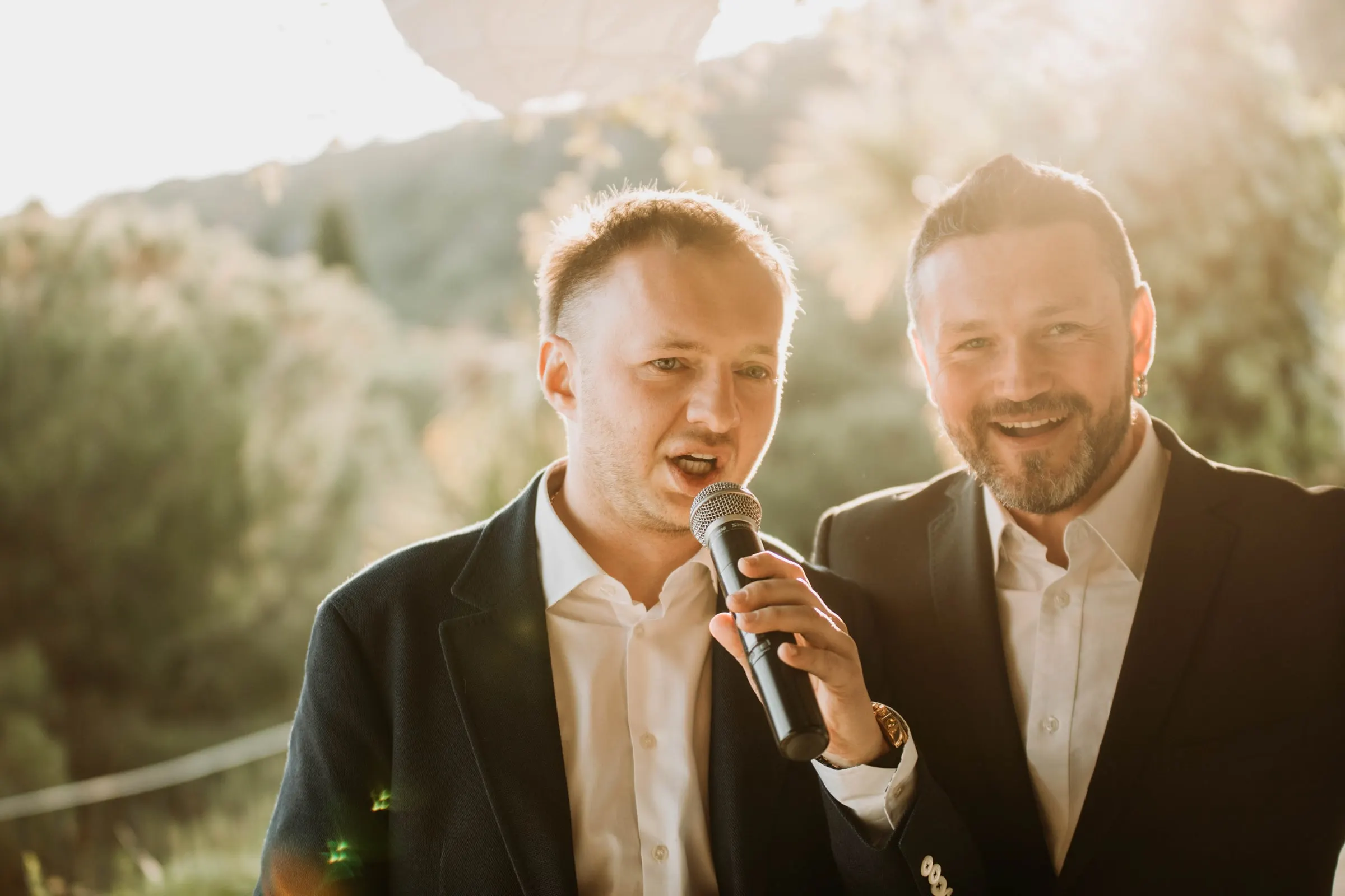 The groom and a friend singing into a microphone together during the toasts at a masia in Girona