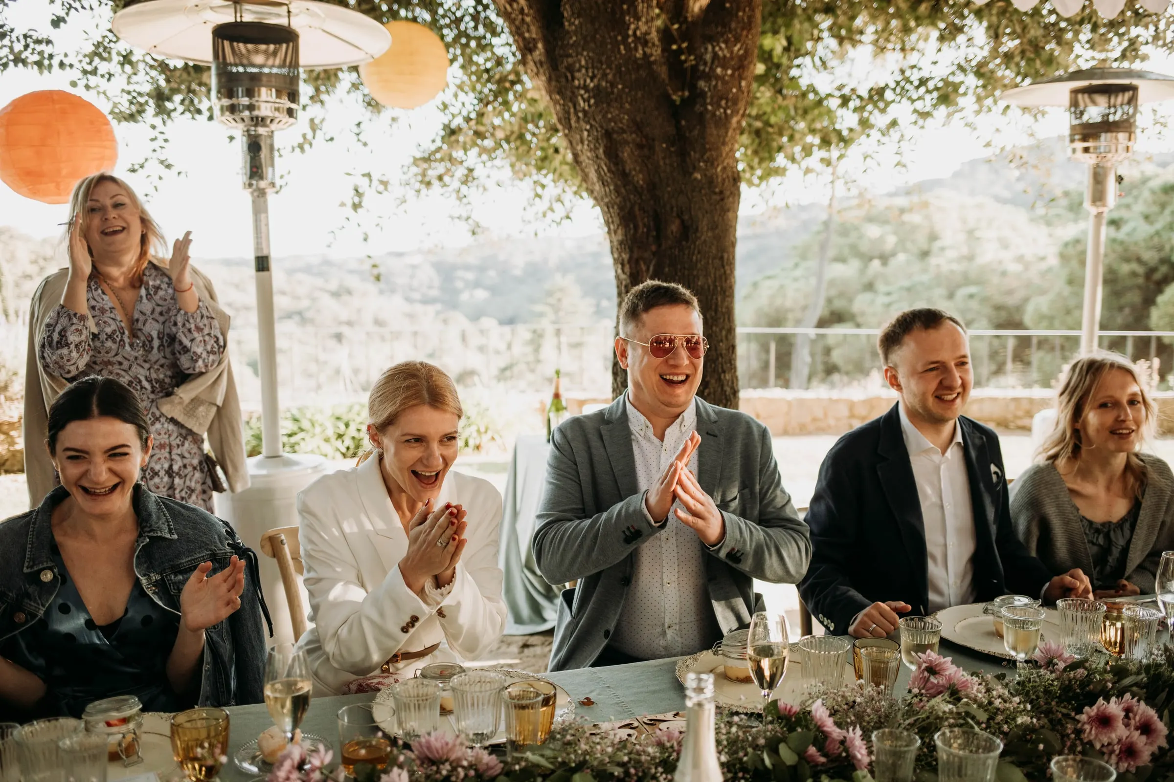 The bride and guests clapping and laughing at the head table under the tree, at a masia in Girona