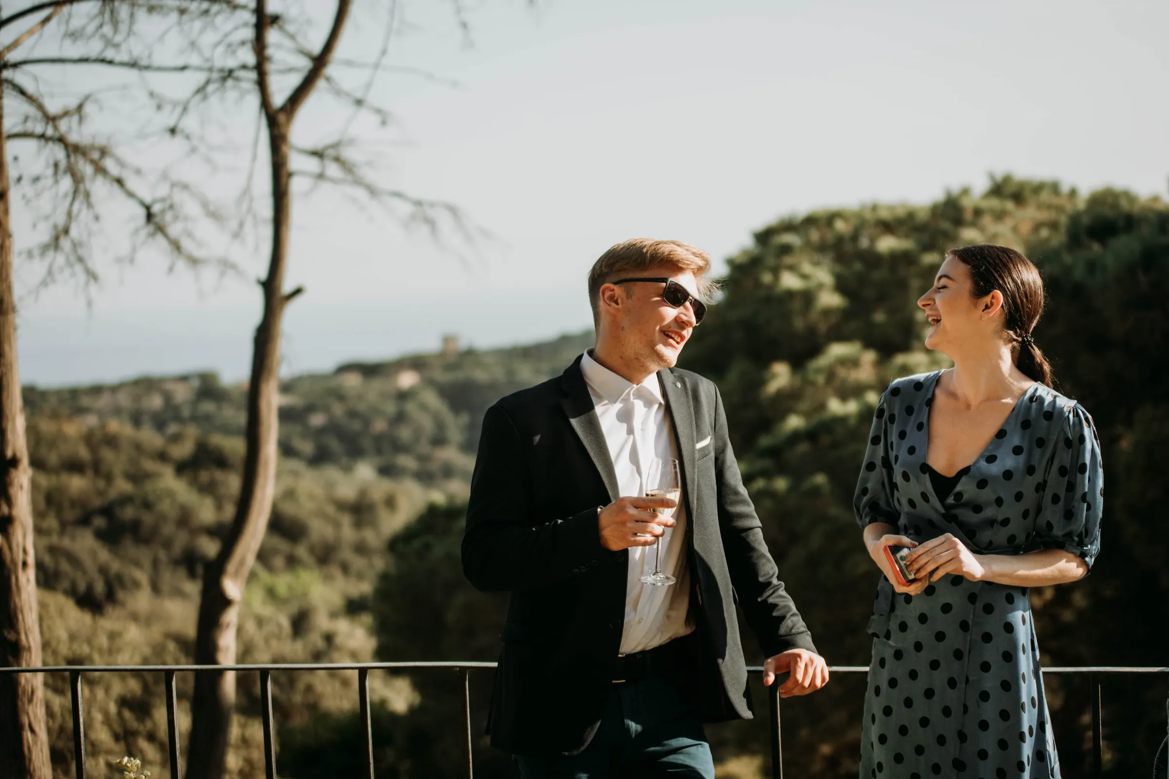 Two guests chatting at the railing with a view over the wooded hills of a masia in Girona