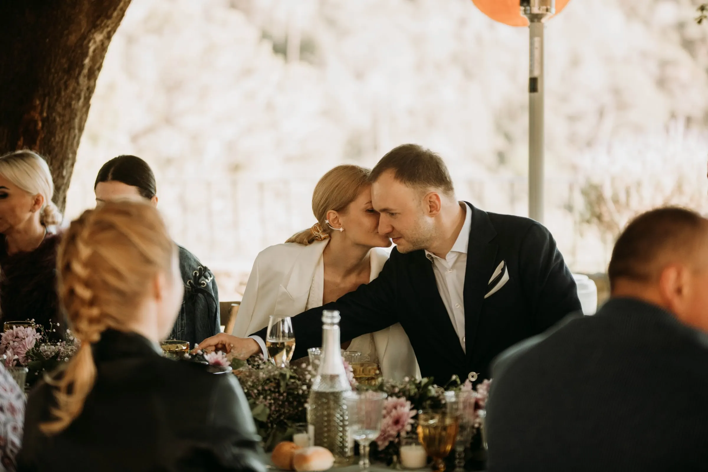 The groom leaning in to kiss the bride at the lunch table among guests, at a masia in Girona