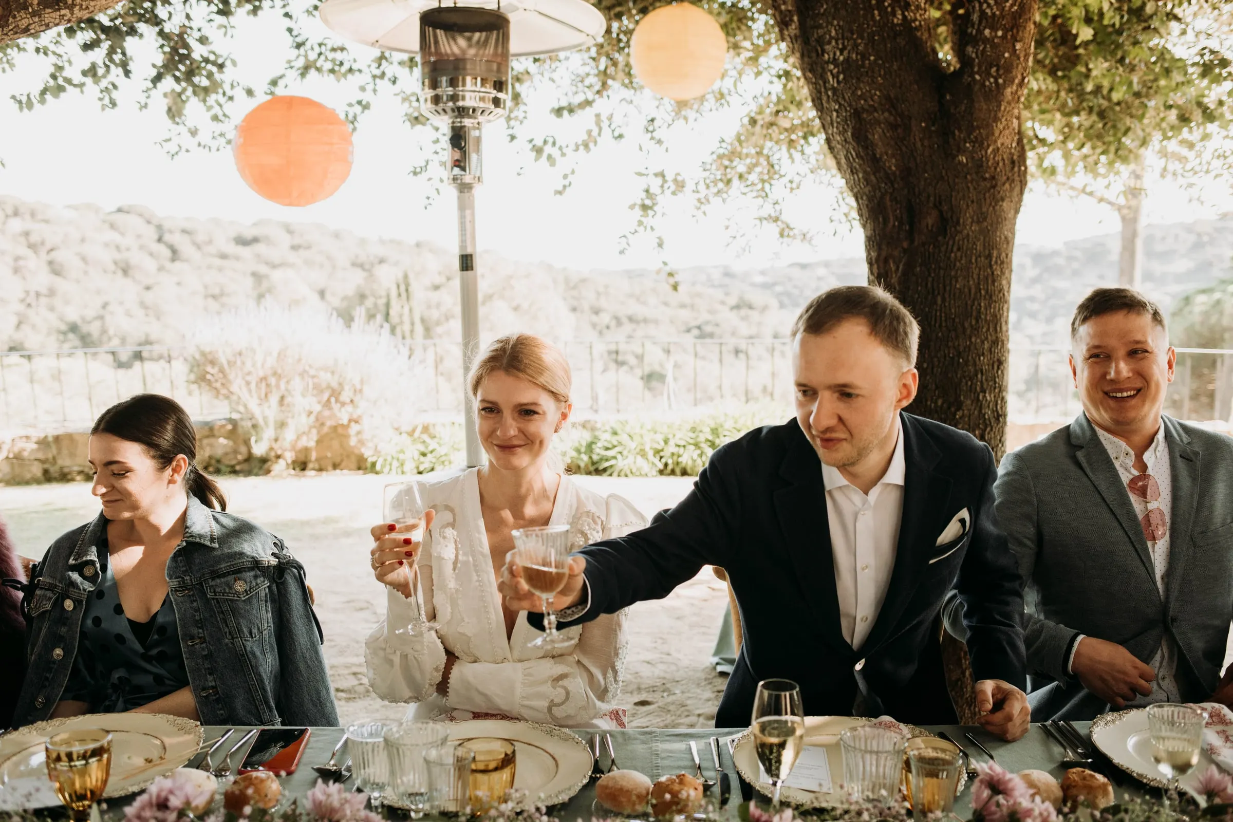 Bride and groom raising their glasses with friends at the table under the tree, at a masia in Girona