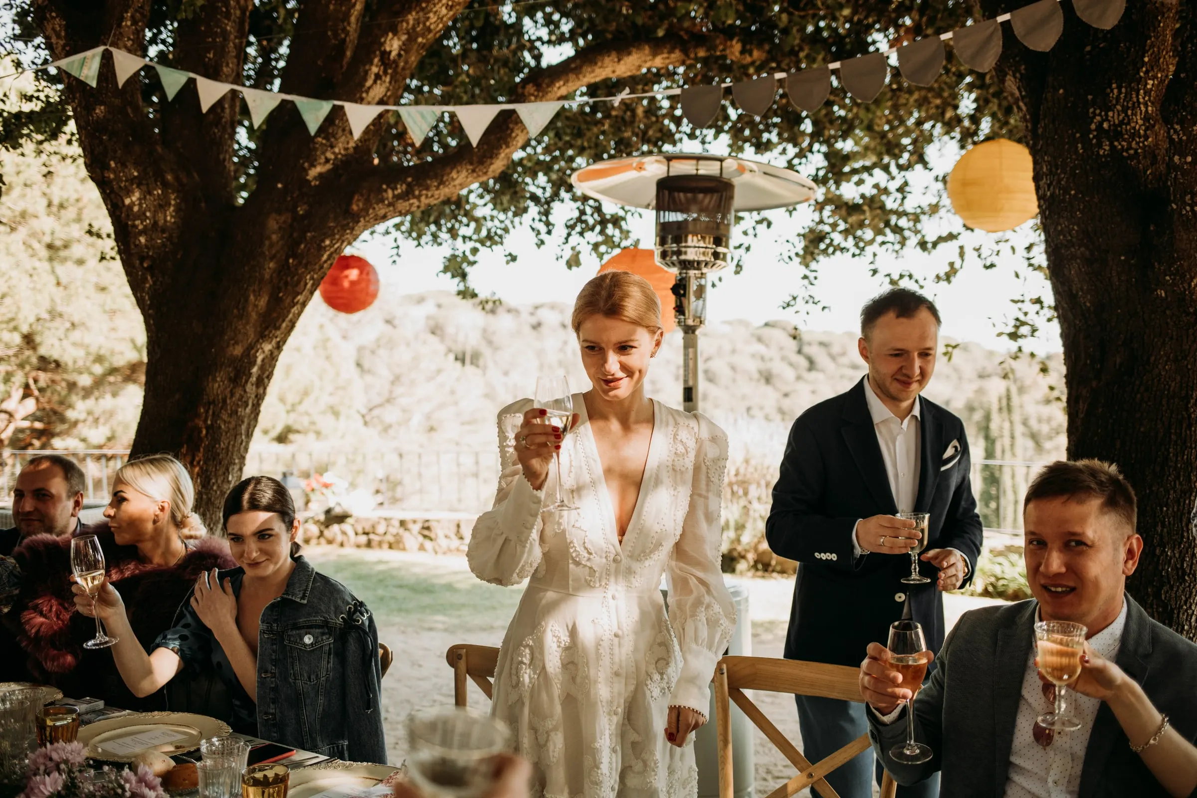 The bride standing to raise a toast as guests cheer under paper lanterns, at a masia in Girona