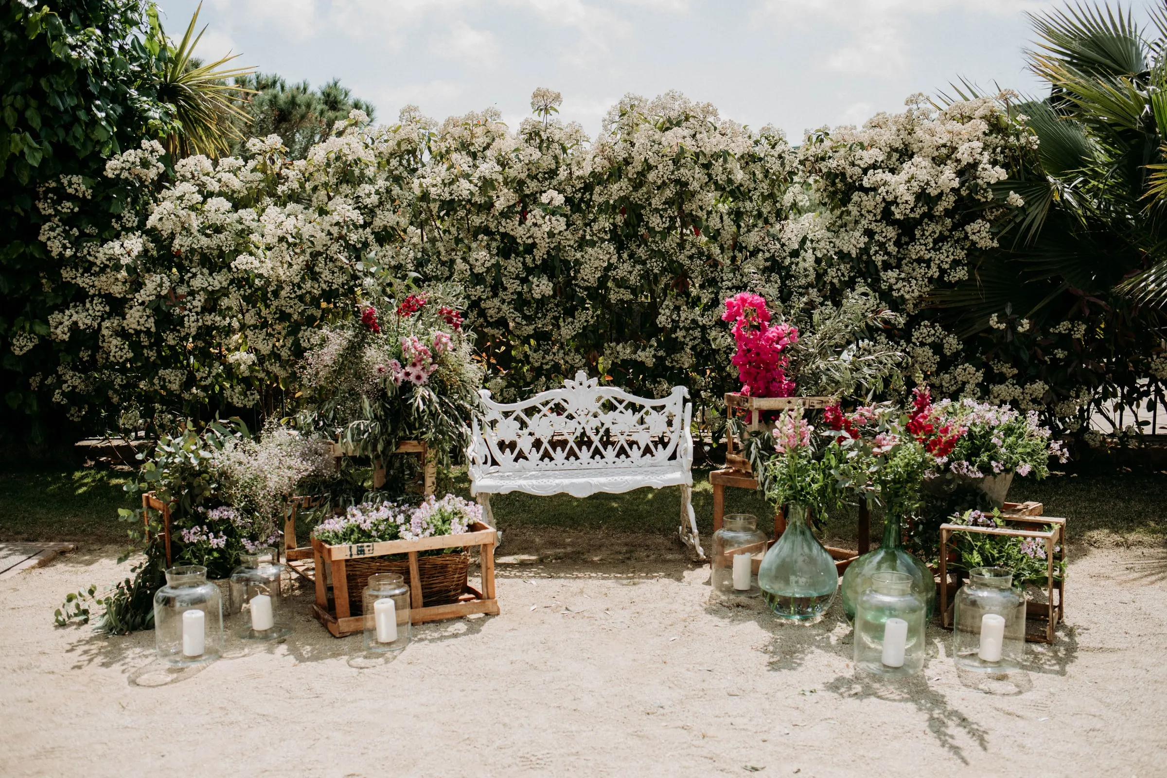 A white iron bench framed by pink and red flowers and glass jars against a blossom hedge, at a masia in Girona